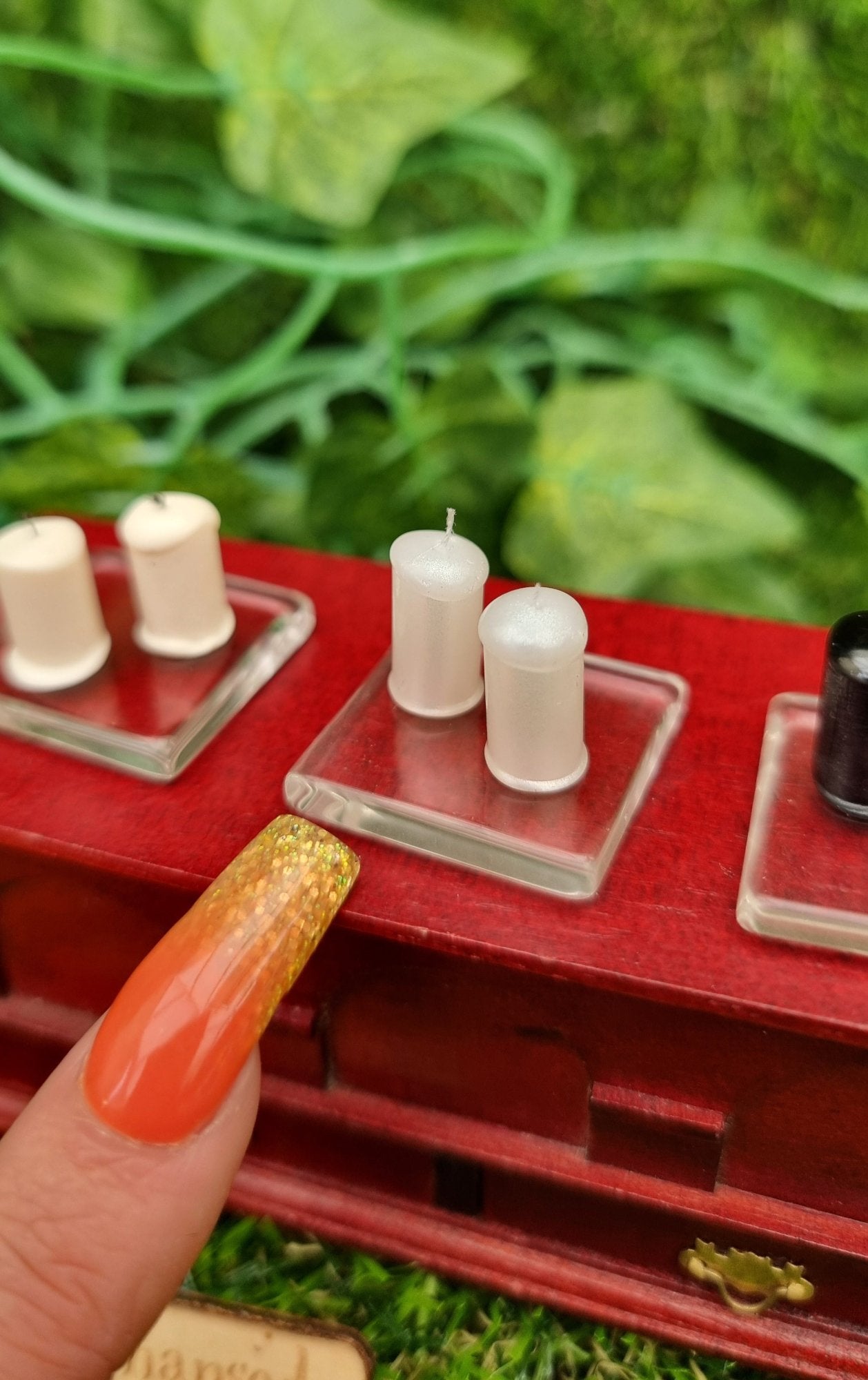 A finger points at 1:12 scale miniature pearlescent white candles sitting on a square glass plate on top of a wooden chest. There are 2 pearlescent white candles with white wicks on the glass plate. @TerraRhapsody is written on wood in the foreground