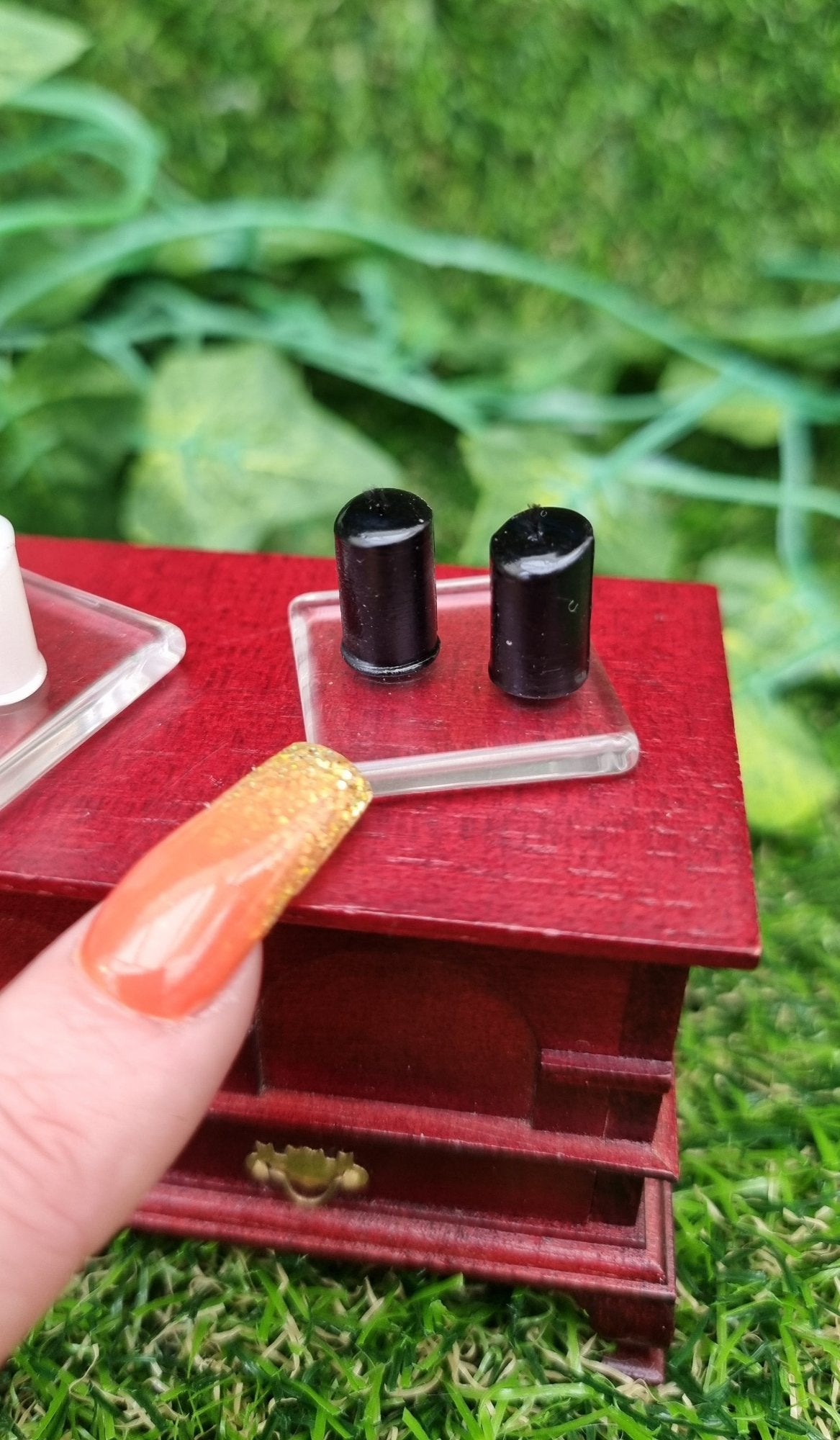 A finger points at 1:12 scale miniature black candles sitting on a square glass plate on top of a wooden chest. There are 2 black candles on the glass plate . @Terra Rhapsody is written on wood in the backgroun