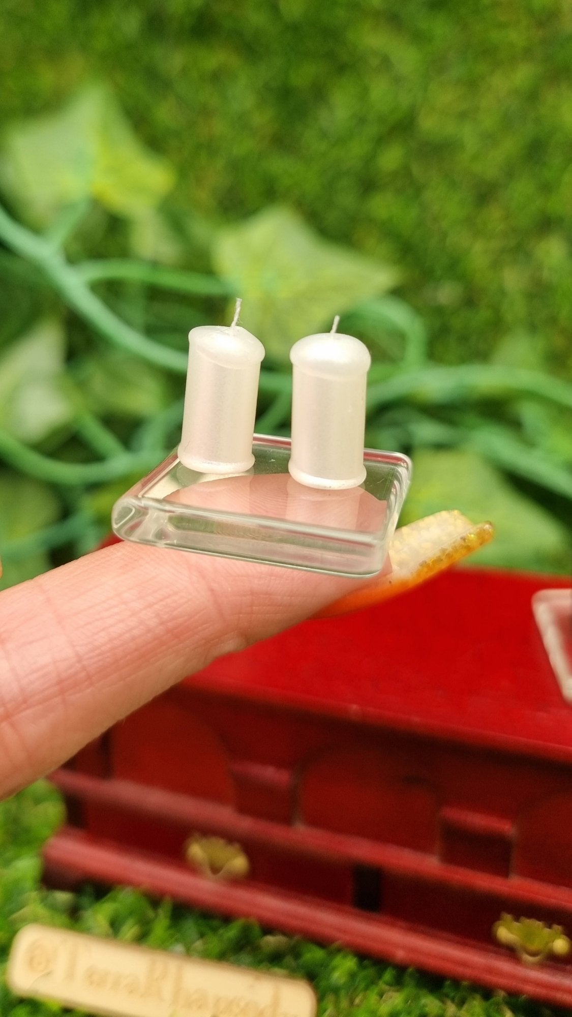 1:12 scale miniature pearlescent white candles sit on a square glass plate on a fingertip in front of a wooden chest. There are 2 candles with white wicks on the glass plate. @TerraRhapsody is written on wood in the background