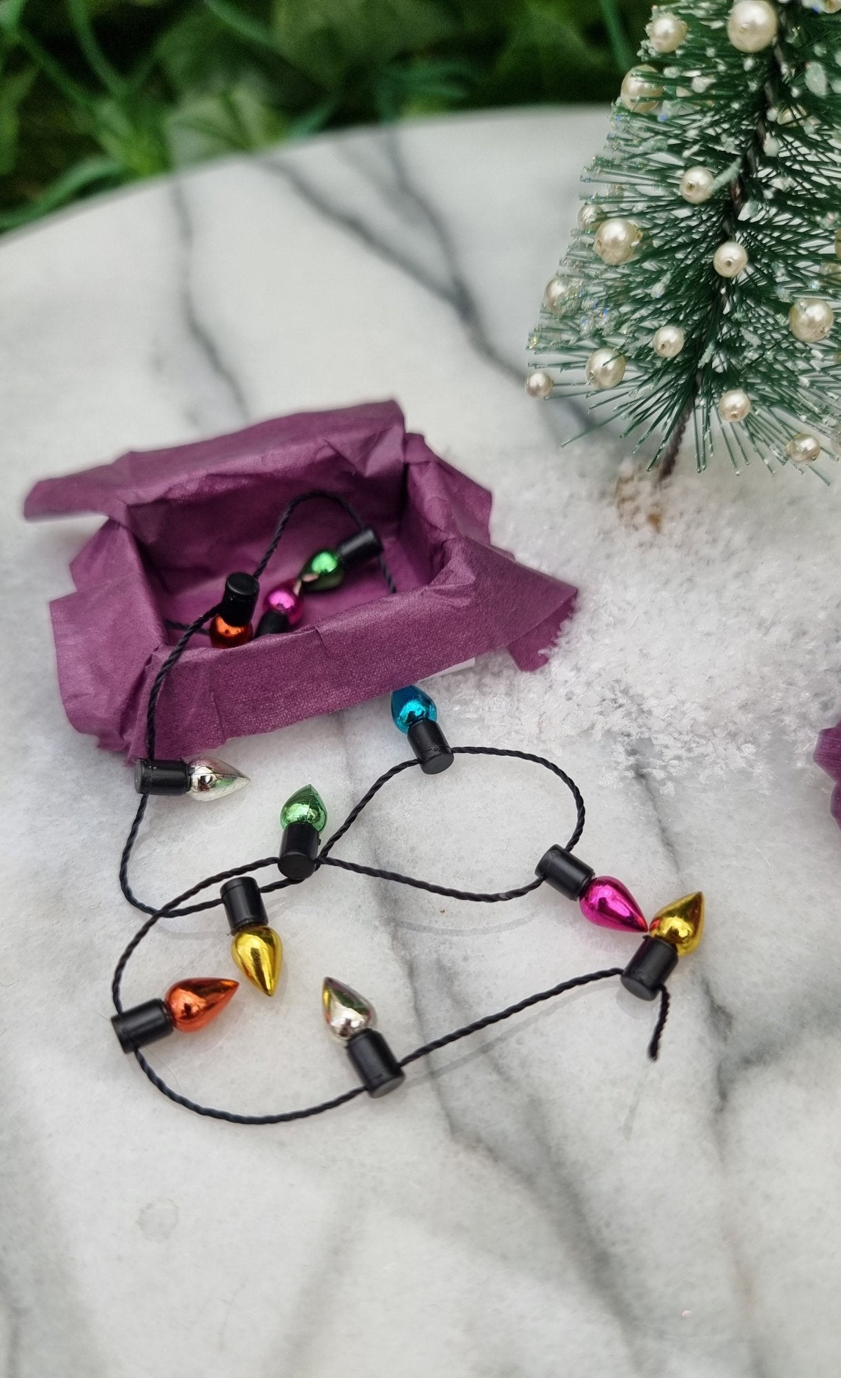 A 1:12 scale miniature box with a string of christmas lights sits on a marble slab. The box is lined with purple paper and the trail of multicoloured lights spill from the box onto the marble