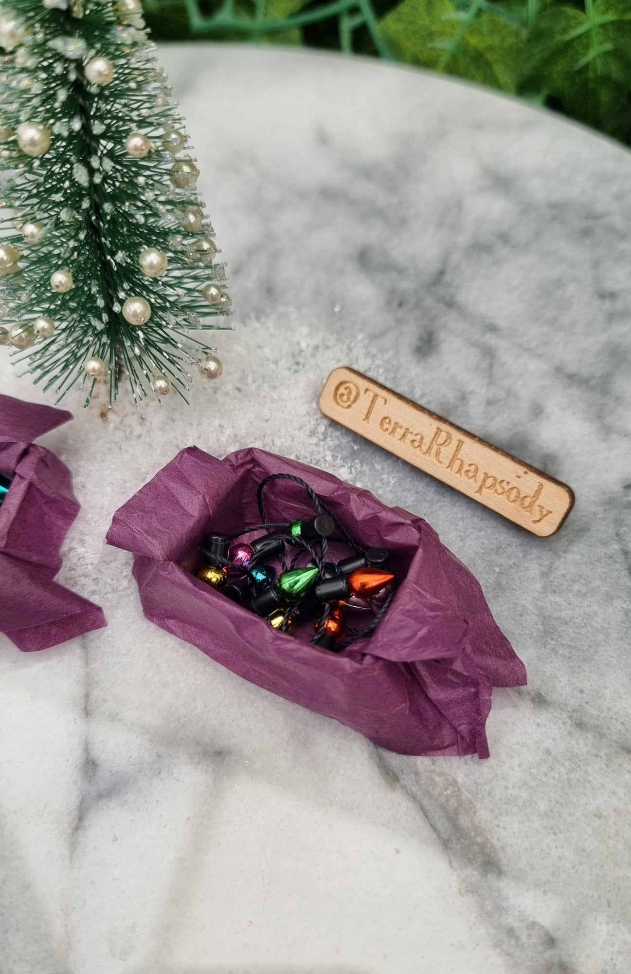 A 1:12 scale miniature box of christmas lights sits on a marble slab. The box is lined with purple paper and contains a string of multicoloured lights. @TerraRhapsody is written on wood beside it