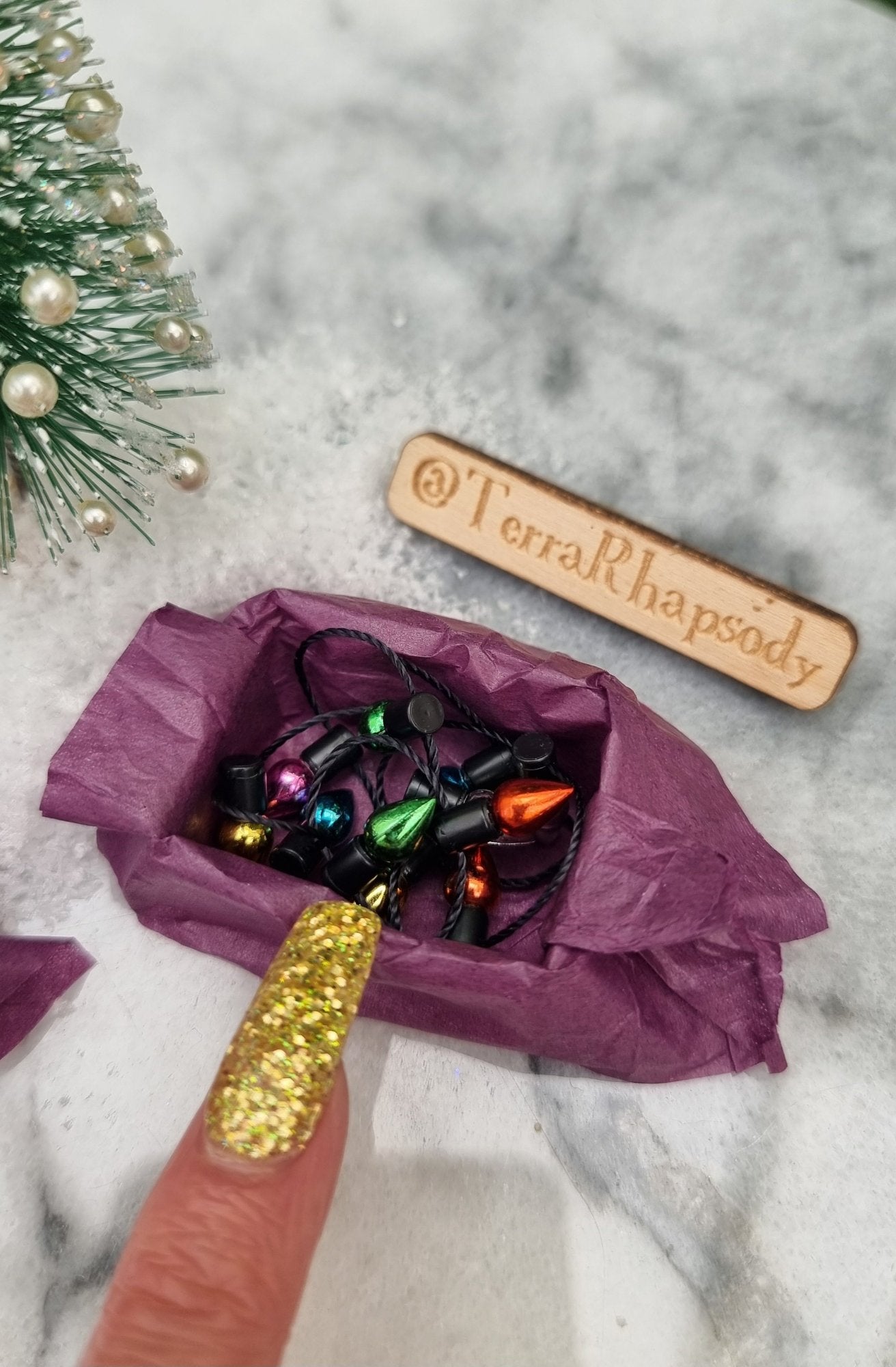 A 1:12 scale miniature box of christmas lights sits on a marble slab. The box is lined with purple paper and contains a string of multicoloured lights. A finger points for scale. @TerraRhapsody is written on wood beside it