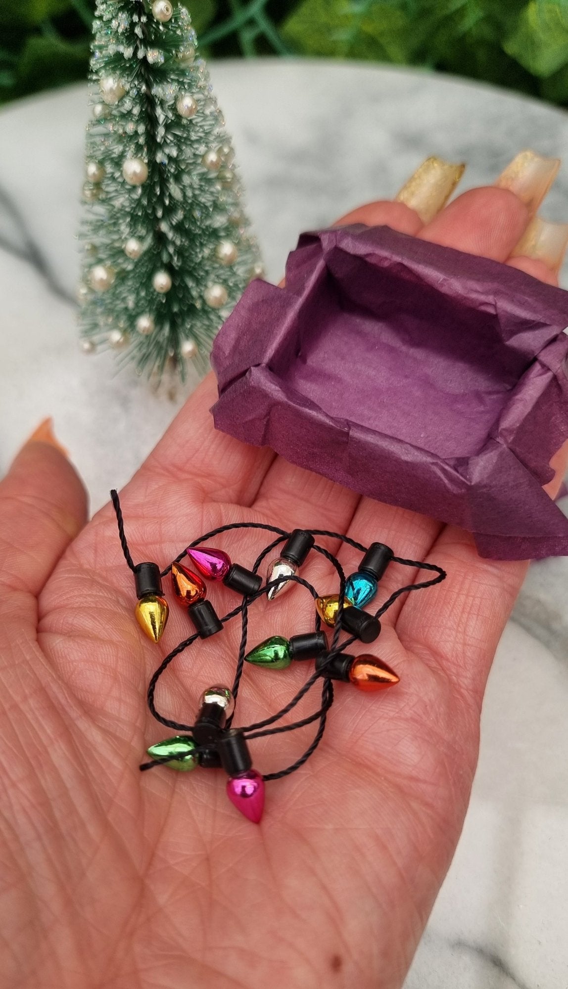 A 1:12 scale miniature box and a string of christmas lights beside it sits in the palm of a hand. The box is lined with purple paper and the trail of multicoloured lights sit in front