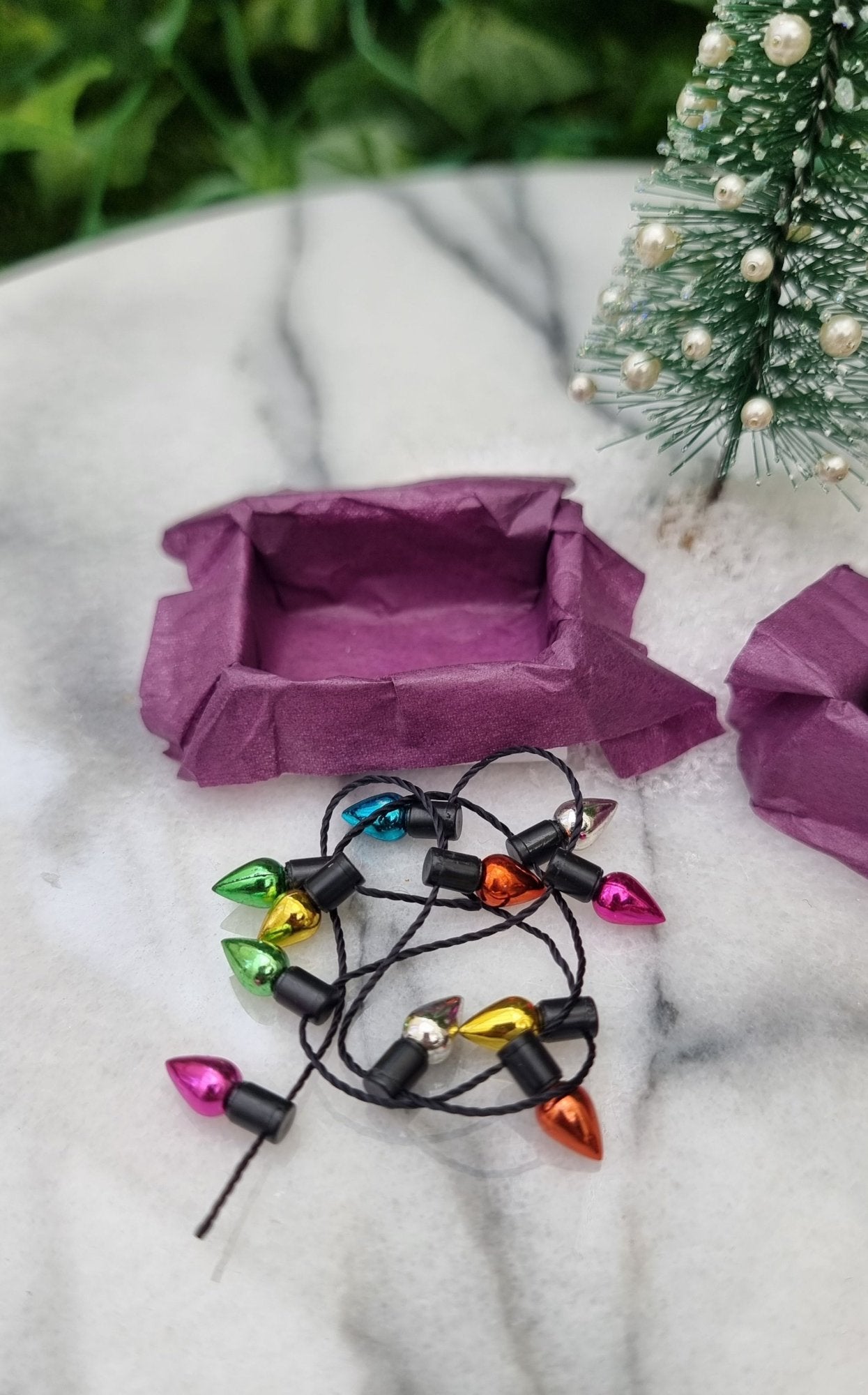 A 1:12 scale miniature box and a string of christmas lights beside it sits on a marble slab. The box is lined with purple paper and the trail of multicoloured lights sit in front