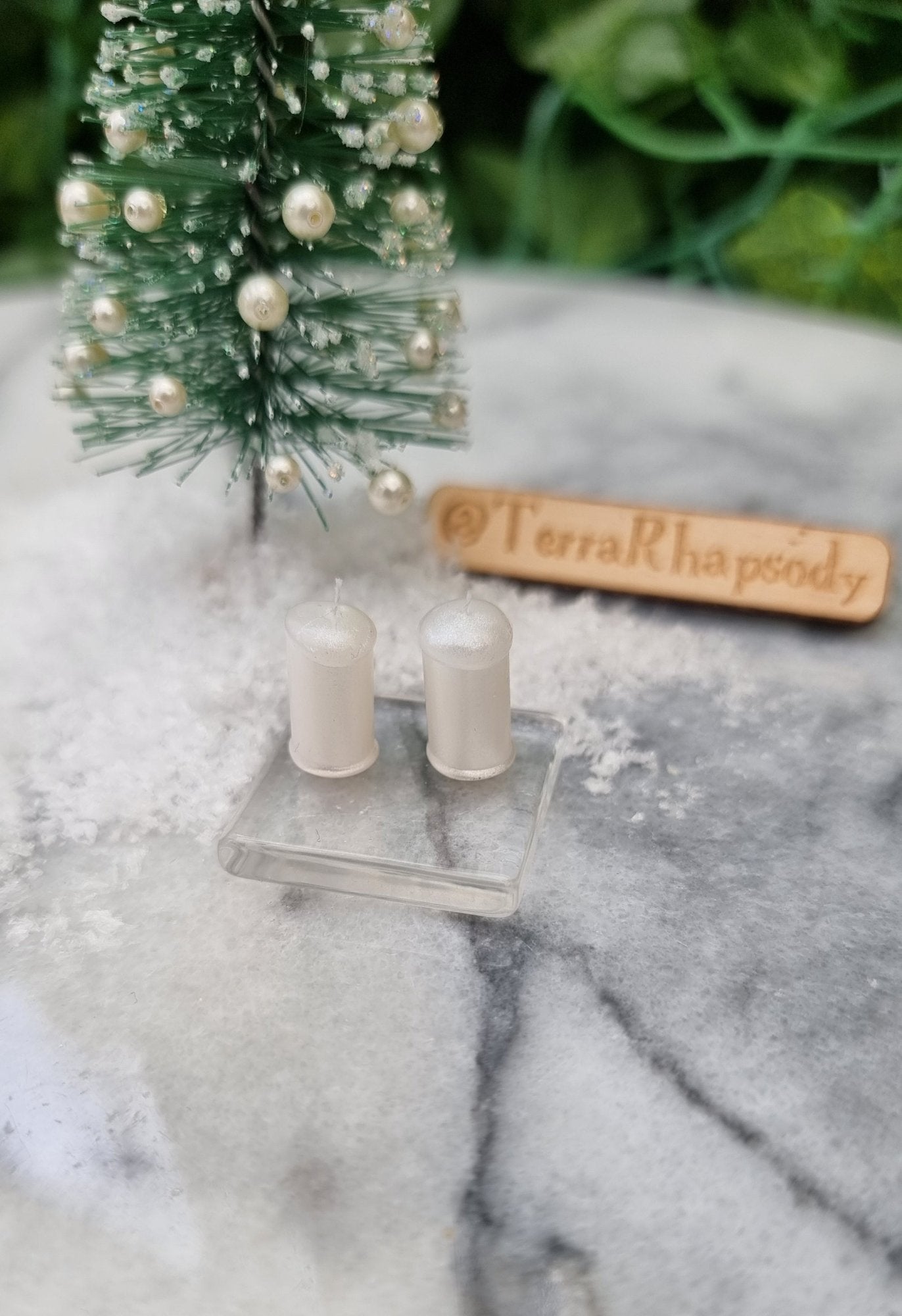 1:12 scale miniature pearlescent white candles sit on a square glass plate in front of a mini christmas tree surrounded by snow. There are 2 candles with white wicks on the glass plate. @TerraRhapsody is written on wood in the background