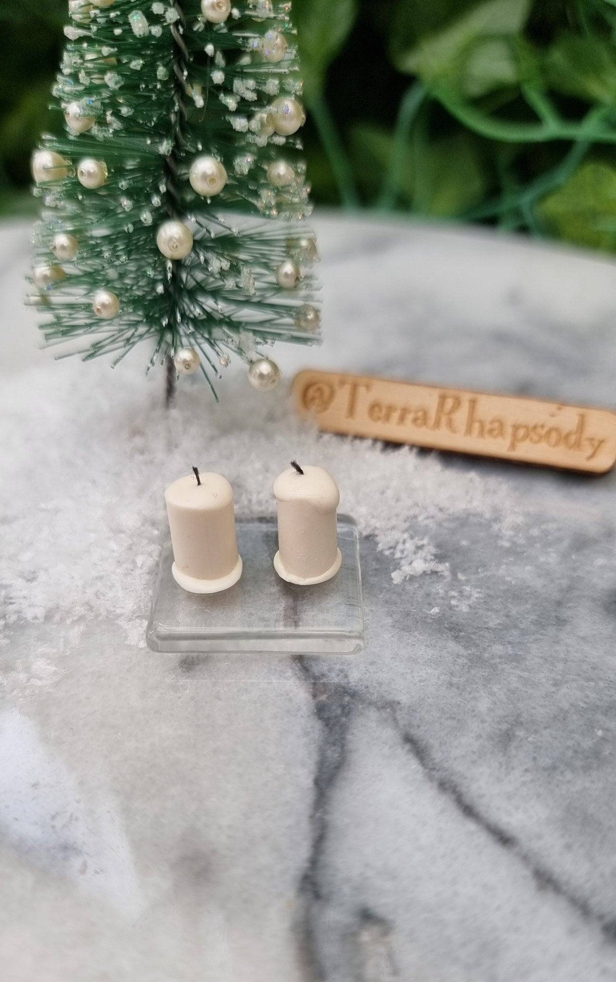 1:12 scale miniature ivory candles sit on a square glass plate in front of a mini christmas tree surrounded by snow. There are 2 ivory candles with black wicks on a the glass plate . @TerraRhapsody is written on wood in the background