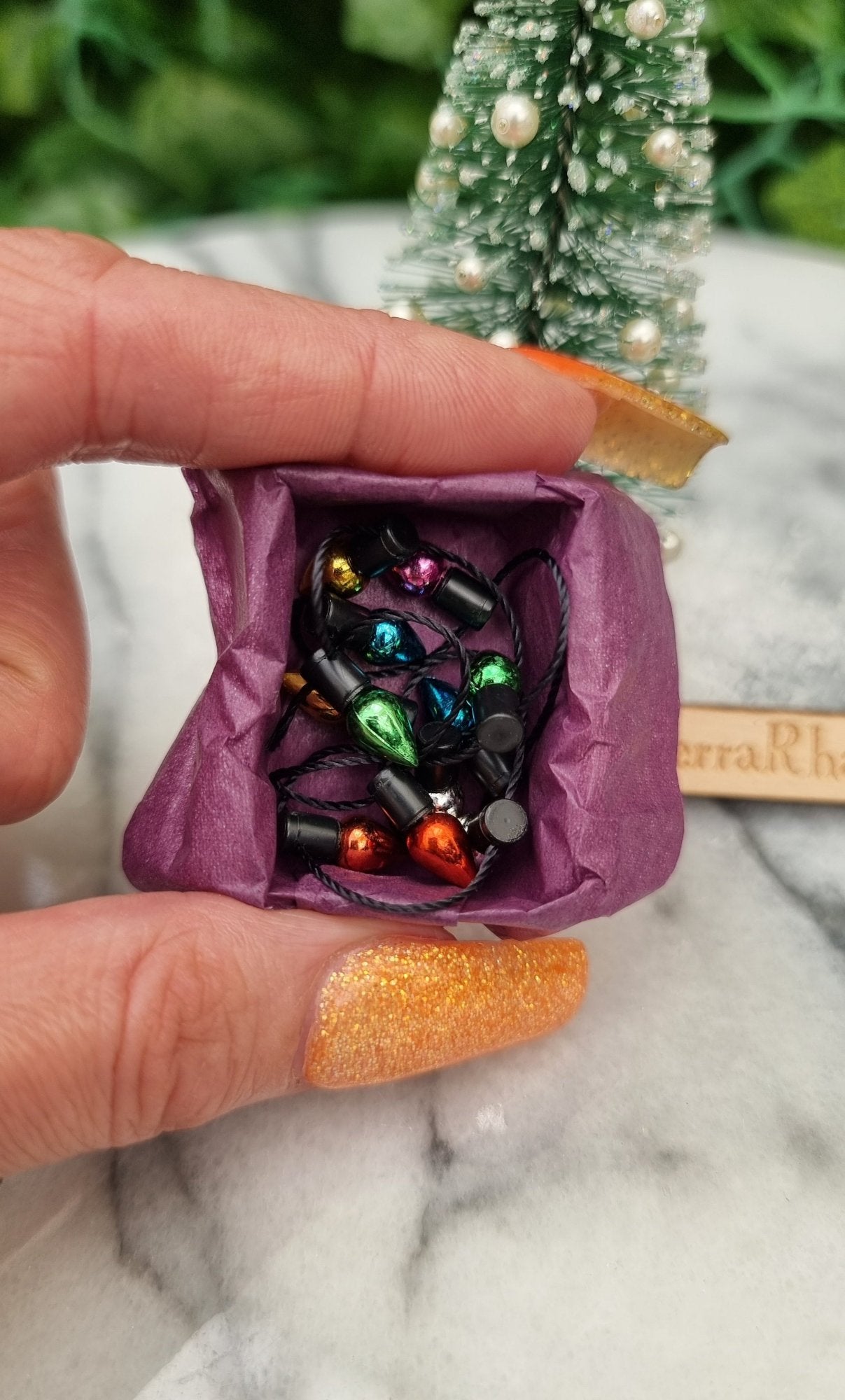 Hand holds a 1:12 scale miniature box of Christmas lights. The box is lined with purple paper filled with multicoloured lights on black string. @TerraRhapsody is written on wood behind it