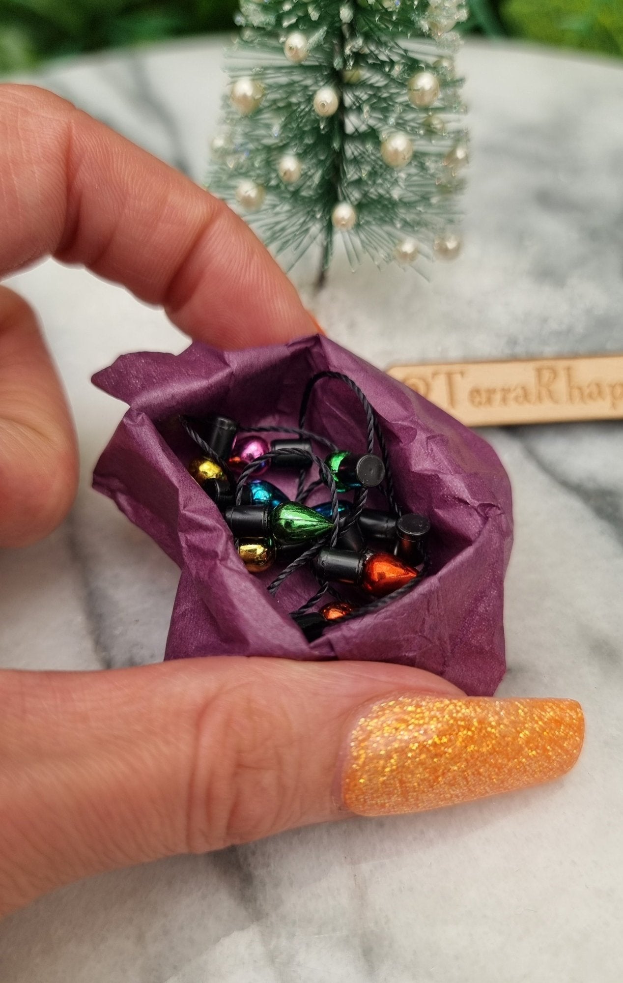 Hand holds a  1:12 scale miniature box of Christmas lights. The box is lined with purple paper filled with multicoloured lights on black string. @TerraRhapsody is written on wood behind it