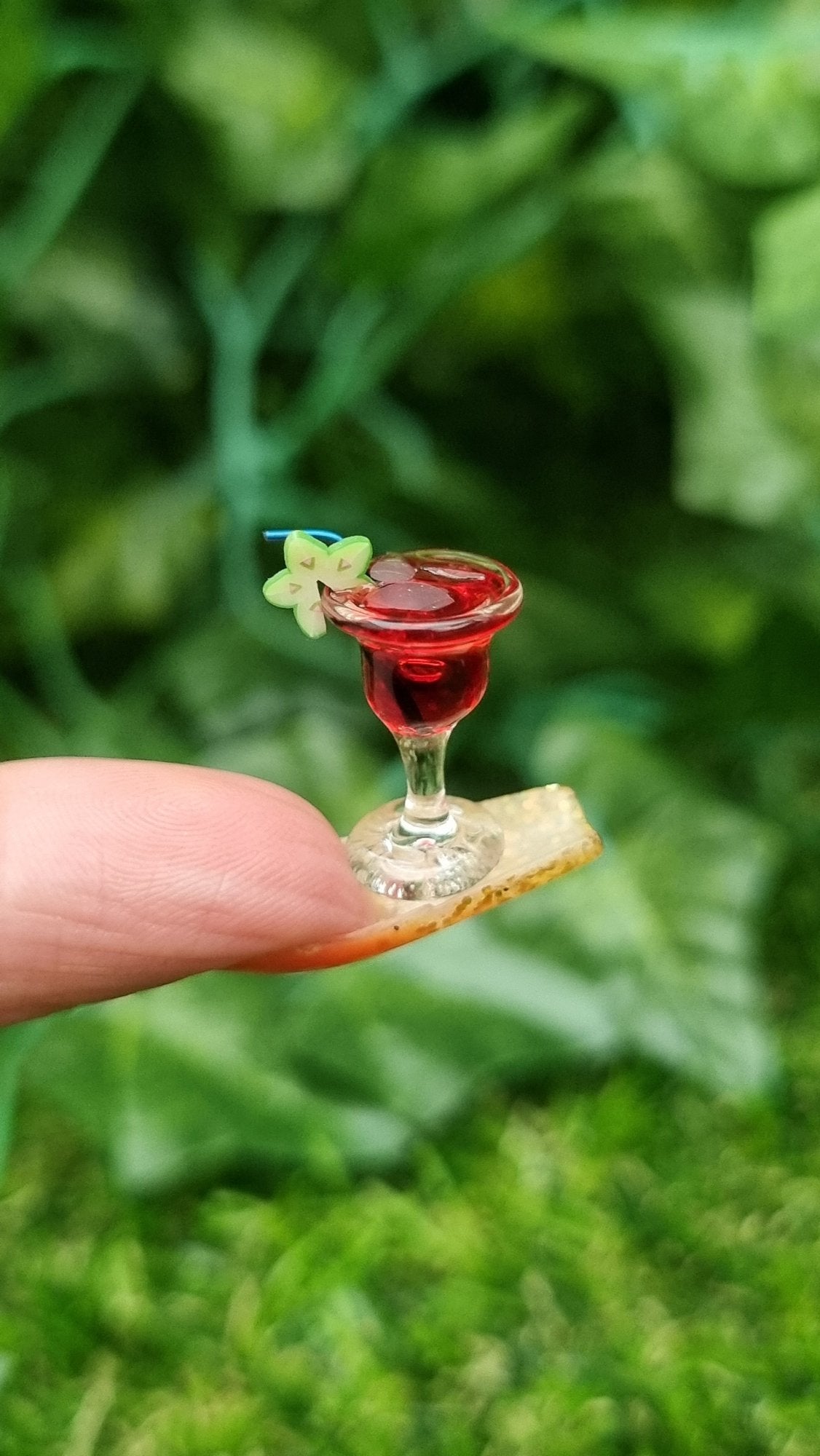 A 1:12 scale miniature cocktail glass is held on the end of a long fingernail. The glass contains red liquid (strawberry daiquiri), turquoise straw and a starfruit slice. Ice is seen on the surface of the liquid