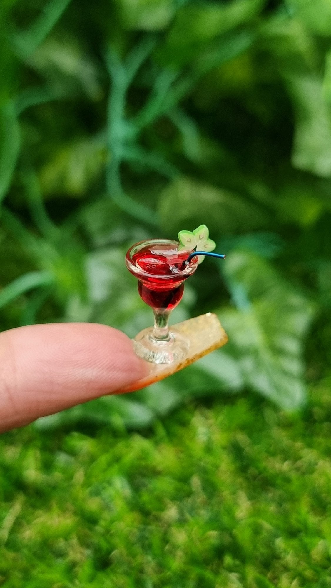 A 1:12 scale miniature cocktail glass is held on the end of a long fingernail. The glass contains red liquid (strawberry daiquiri), turquoise straw and a starfruit slice. Ice is seen on the surface of the liquid