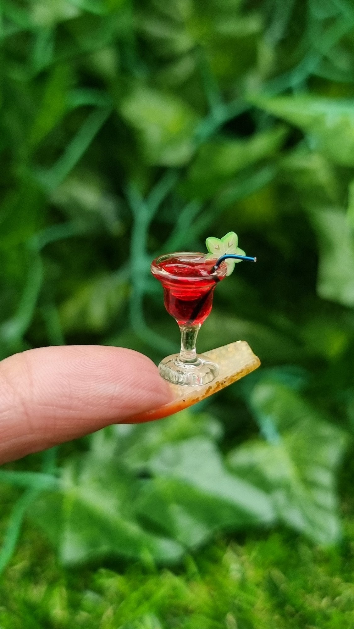 A 1:12 scale miniature cocktail glass is held on the end of a long fingernail. The glass contains red liquid (strawberry daiquiri), turquoise straw and a starfruit slice. Ice is seen on the surface of the liquid