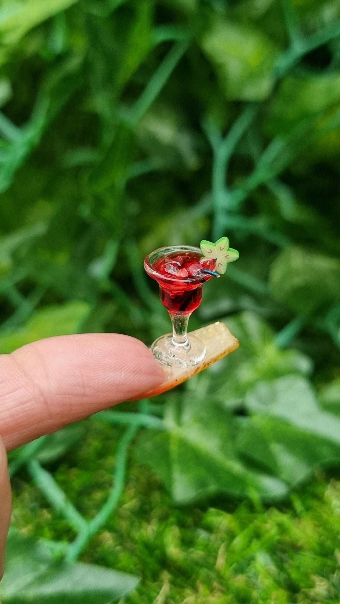 A 1:12 scale miniature cocktail glass is held on the end of a long fingernail. The glass contains red liquid (strawberry daiquiri), turquoise straw and a starfruit slice. Ice is seen on the surface of the liquid