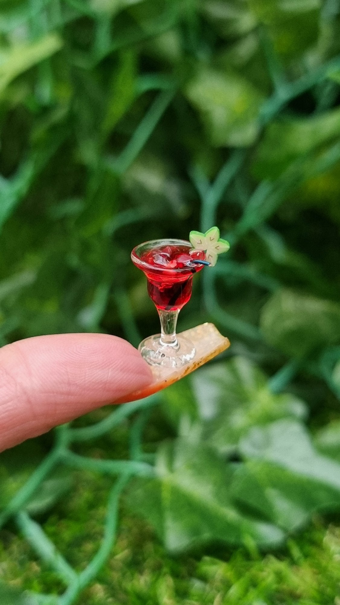 A 1:12 scale miniature cocktail glass is held on the end of a long fingernail. The glass contains red liquid (strawberry daiquiri), turquoise straw and a starfruit slice. Ice is seen on the surface of the liquid