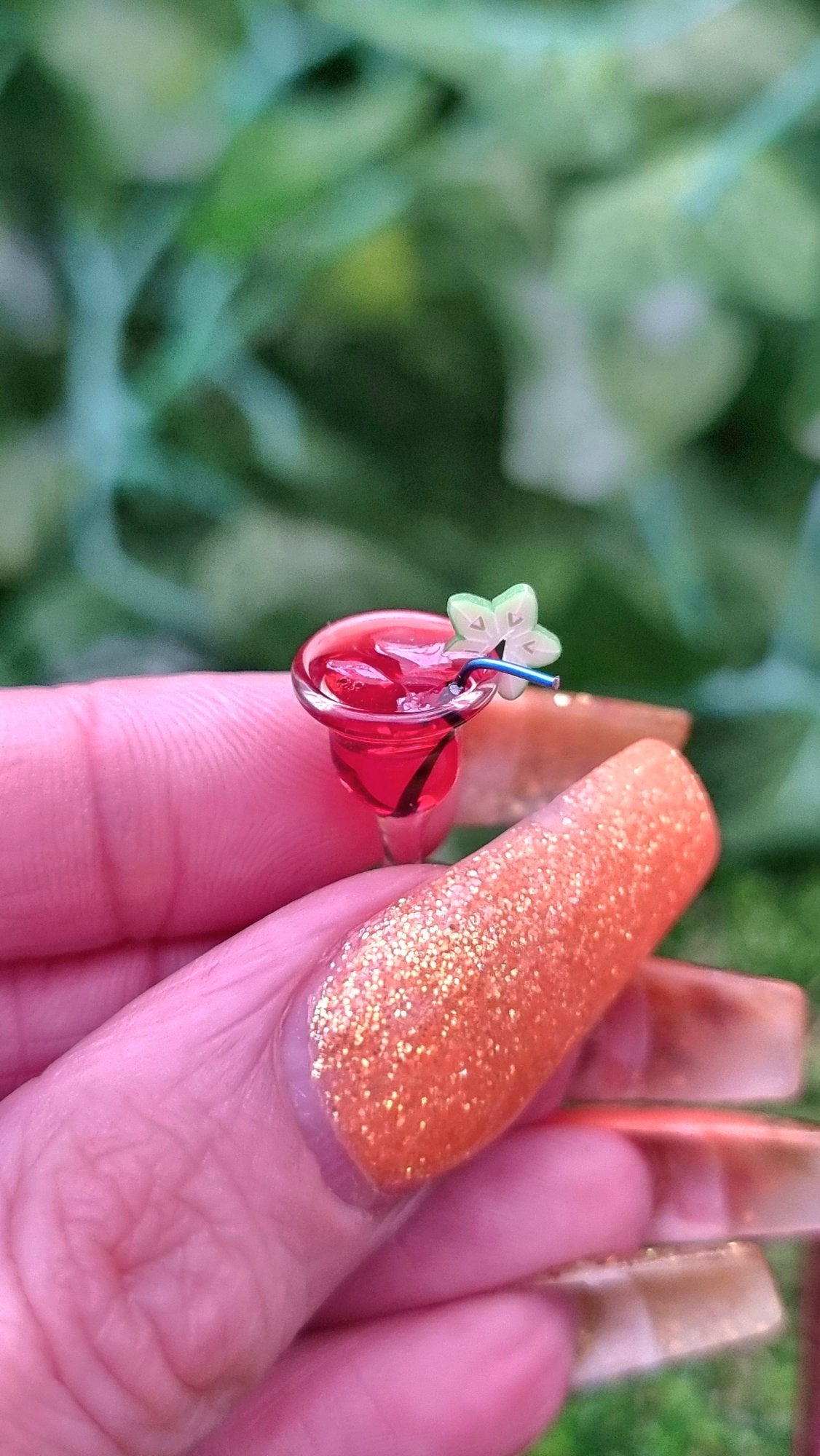 A 1:12 scale miniature cocktail glass is held between a finger and thumb. The glass contains red liquid (strawberry daiquiri), turquoise straw and a starfruit slice. Ice is seen on the surface of the liquid