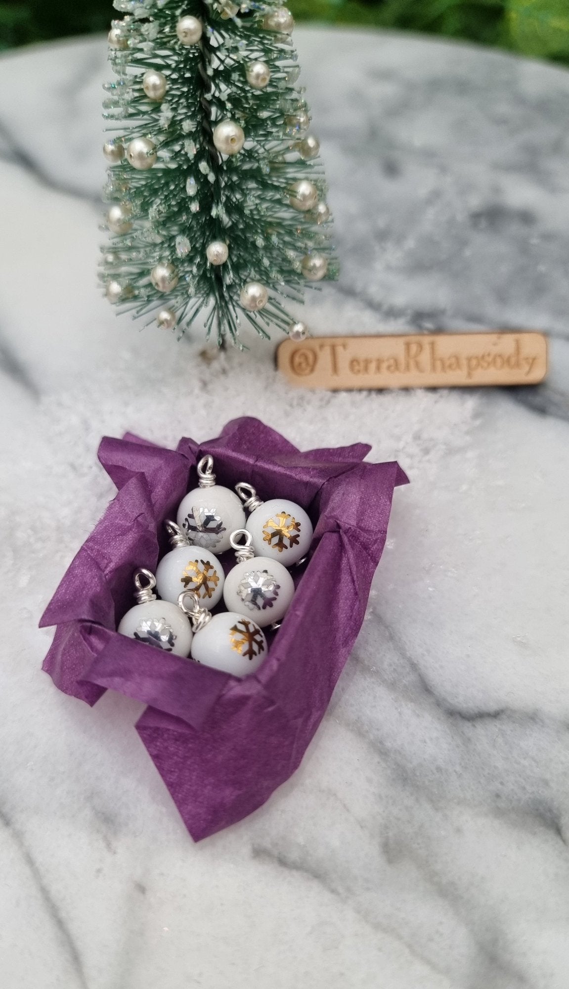 Miniature 1:12 scale open tray box contains 6 Christmas baubles. The baubles are white with gold and silver snowflake motifs. The box is lined with purple paper, displayed on snow and marble. @TerraRhapsody is written on wood in the background