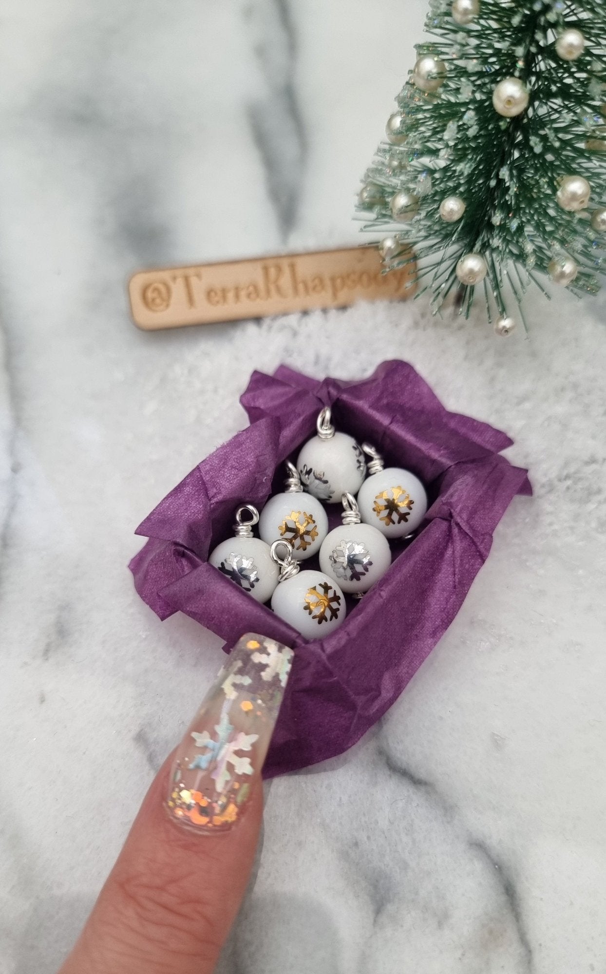 Miniature 1:12 scale open tray box contains 6 Christmas baubles. The baubles are white with gold and silver snowflake motifs. The box is lined with purple paper, displayed on snow and marble. A finger points for scale. @TerraRhapsody is written on wood in the background