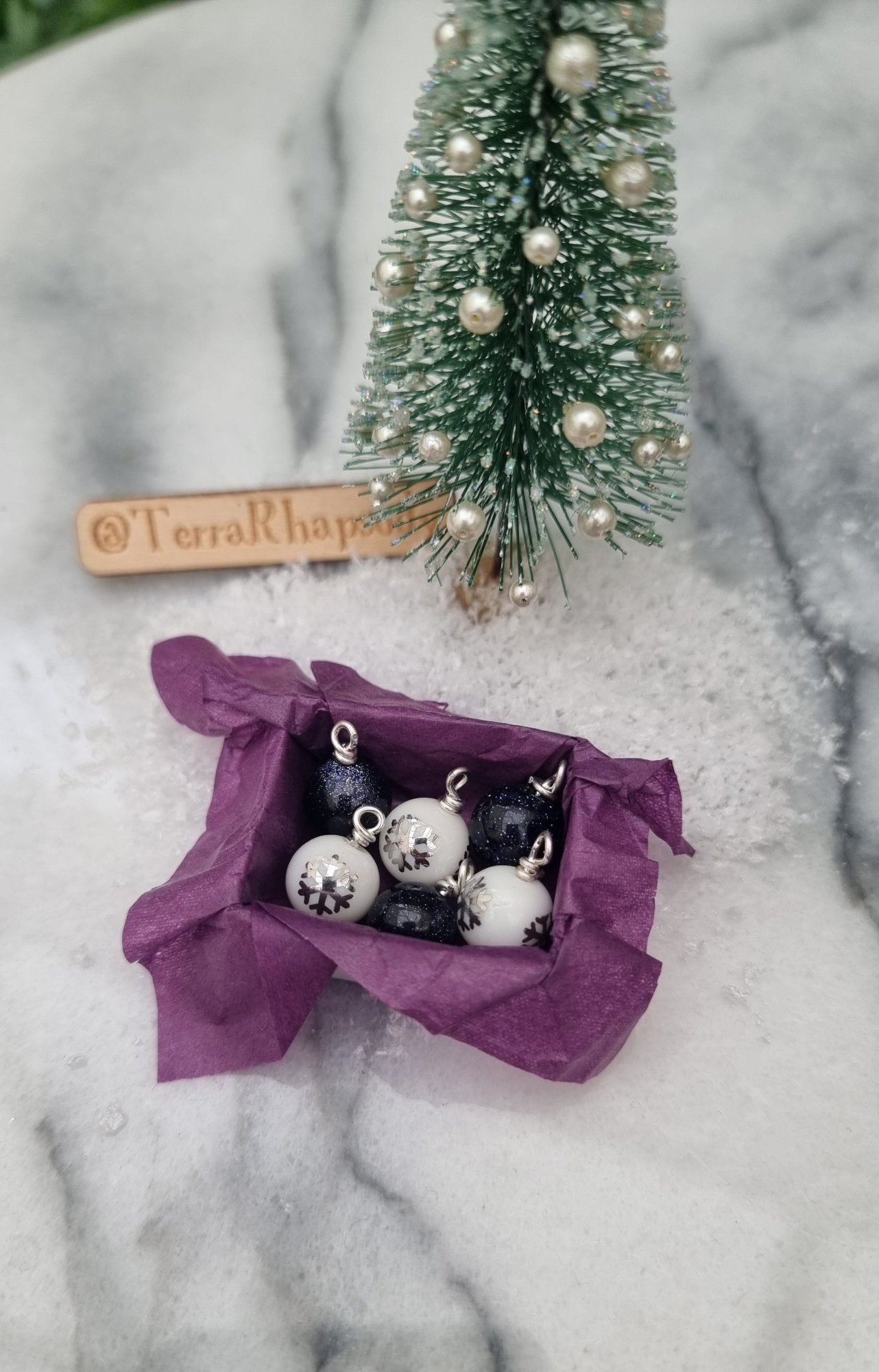 Miniature 1:12 scale open tray box contains 6 Christmas baubles. 3 baubles are white with silver snowflake motifs, 3 are glittery blue goldstone. The box is lined with purple paper. @TerraRhapsody is written on wood in the background