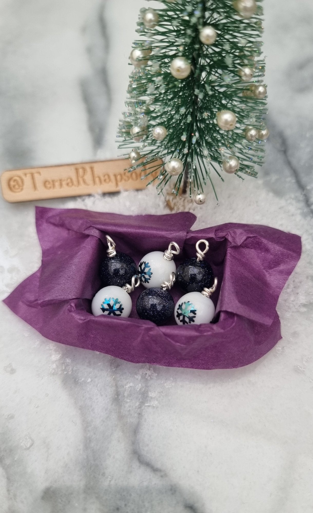 Miniature 1:12 scale open tray box contains 6 Christmas baubles. 3 baubles are white with blue snowflake motifs, 3 are glittery blue goldstone. The box is lined with purple paper. @TerraRhapsody is written on wood in the background