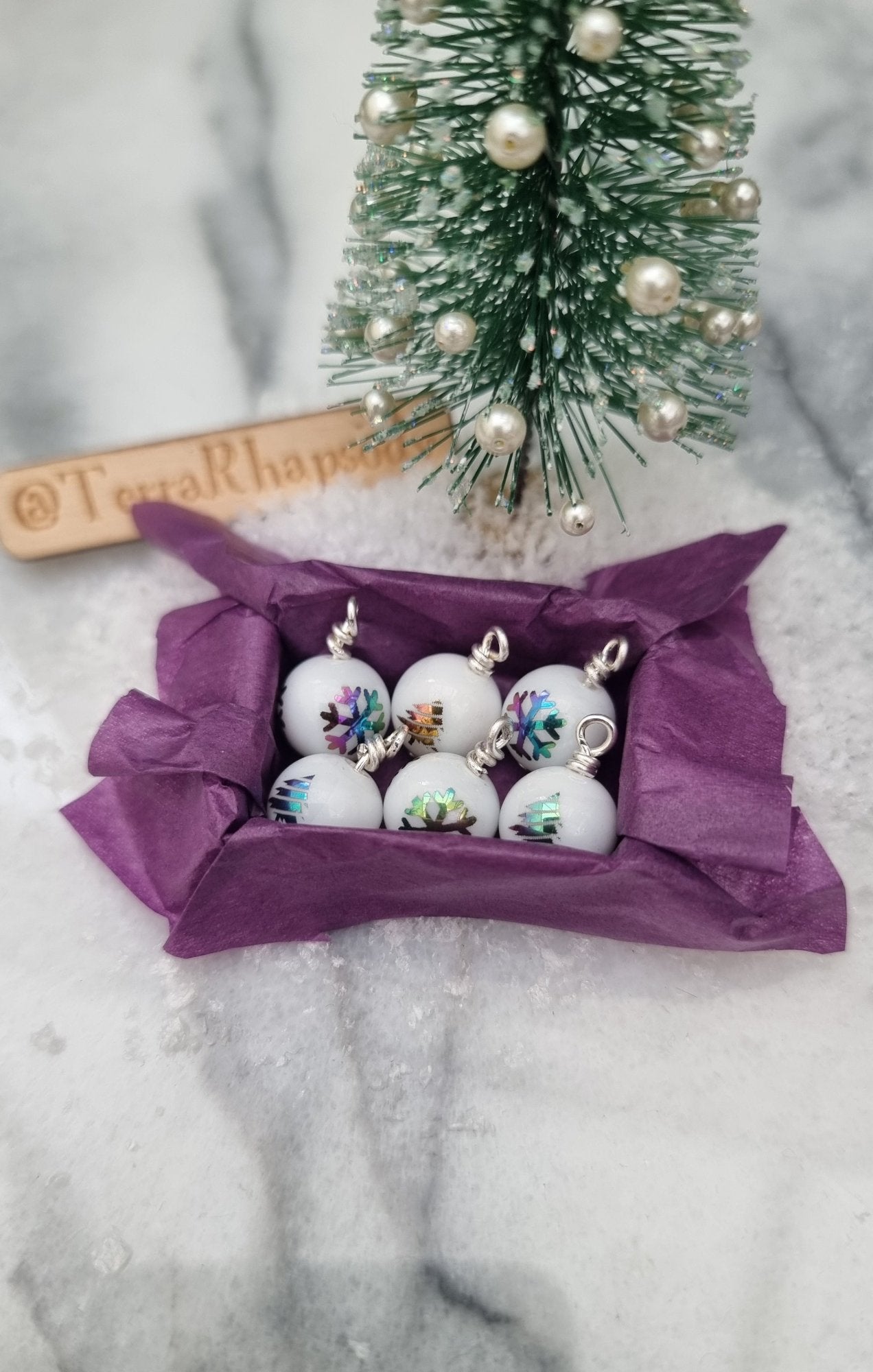 Miniature 1:12 scale open tray box contains 6 Christmas baubles. The baubles are white with multicoloured snowflake and tree motifs. The box is lined with purple paper, displayed on snow and marble. @TerraRhapsody is written on wood in the background