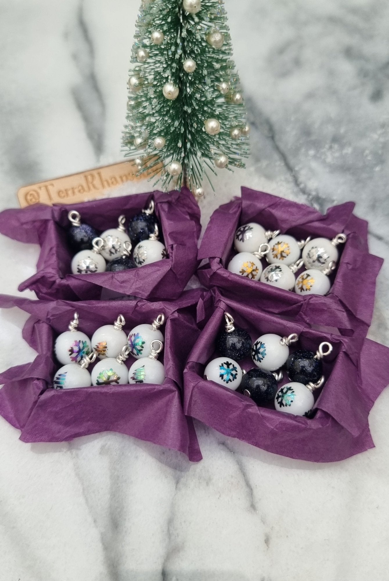 4 miniature 1:12 scale open tray boxes containing 6 Christmas baubles are displayed on a marble slab. The baubles are white with coloured festive motifs and blue goldstone. The boxes are lined with purple paper. @TerraRhapsody is written on wood in the background