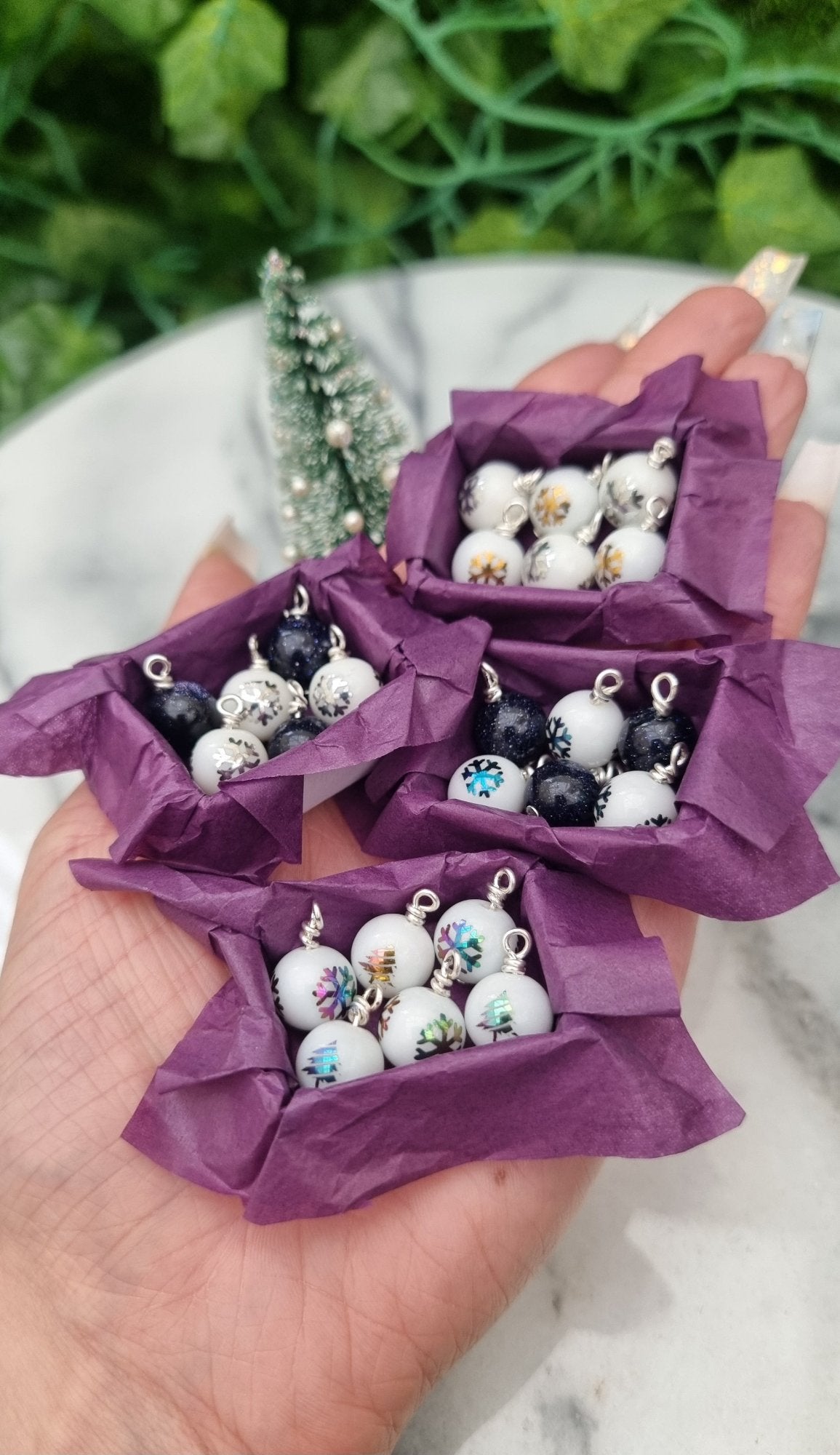 4 miniature 1:12 scale open tray boxes containing 6 Christmas baubles are displayed on a hand. The baubles are white with coloured festive motifs and blue goldstone. The boxes are lined with purple paper. @TerraRhapsody is written on wood in the background