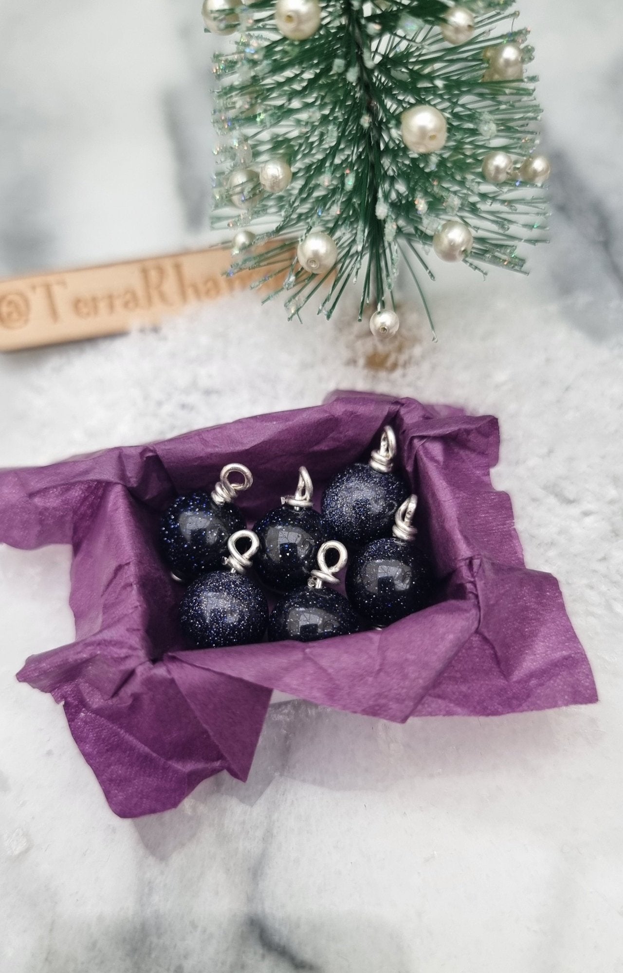 Miniature 1:12 scale open tray box contains 6 Christmas baubles. The baubles are glittering blue goldstone. The box is lined with purple paper, displayed on marble slab. @TerraRhapsody is written on wood in the background