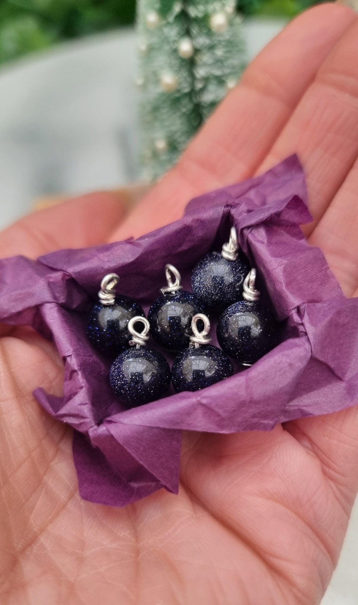 Miniature 1:12 scale open tray box contains 6 Christmas baubles. The baubles are glittering blue goldstone. The box is lined with purple paper, displayed on a hand. @TerraRhapsody is written on wood in the background