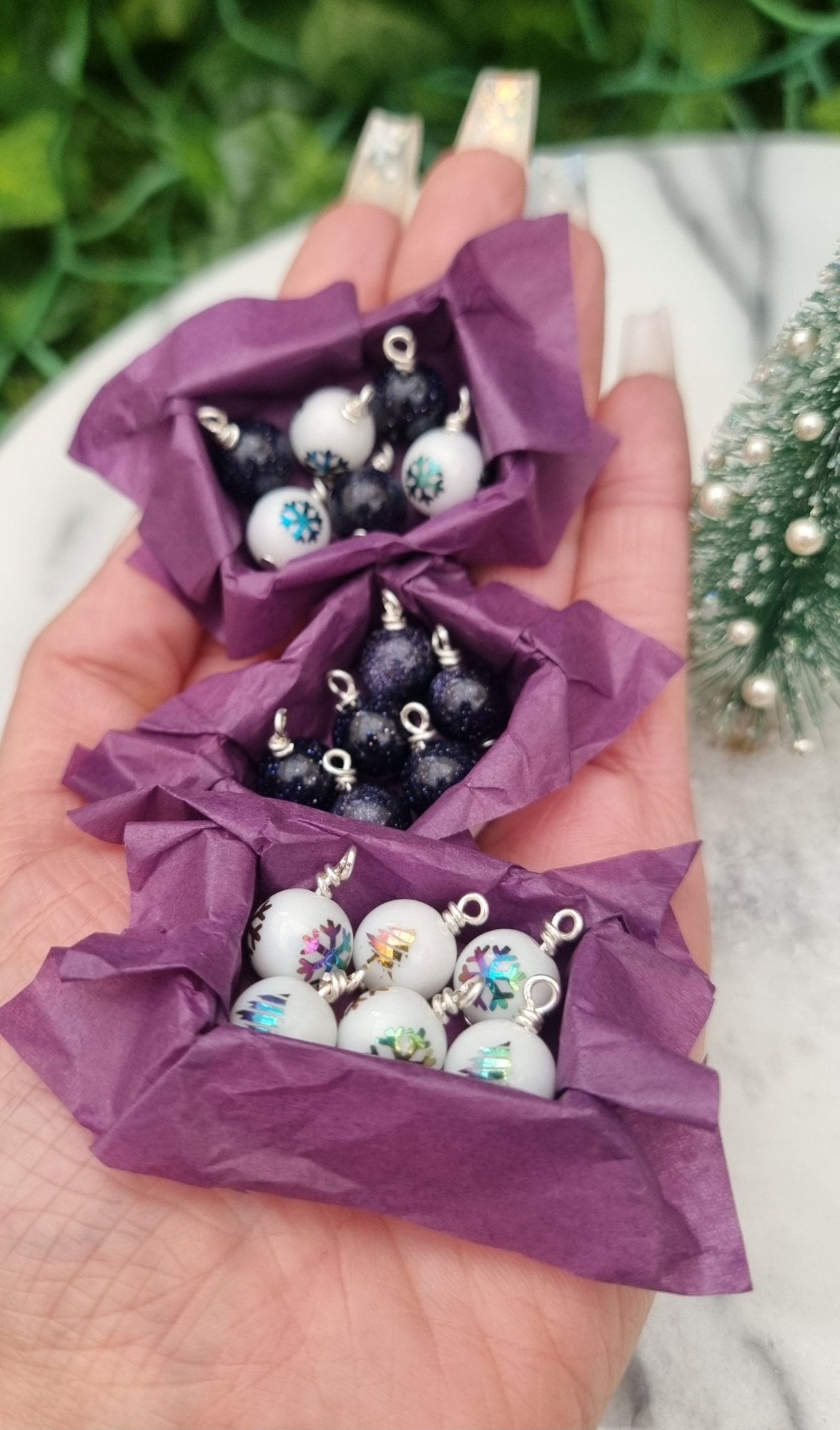 3 miniature 1:12 scale open tray boxes containing 6 Christmas baubles are displayed on a hand. The baubles are white with coloured festive motifs and blue goldstone. The boxes are lined with purple paper. @TerraRhapsody is written on wood in the background