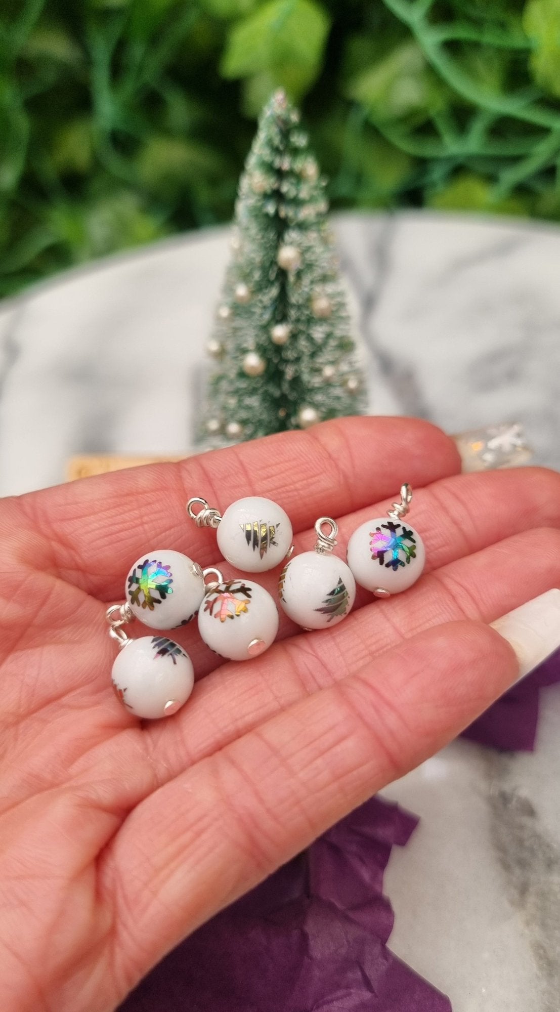 6 Miniature 1:12 Christmas baubles are displayed a hand in front of a Christmas tree. The baubles are white with rainbow coloured snowflake and Christmas tree motifs. @TerraRhapsody is written on wood in the background