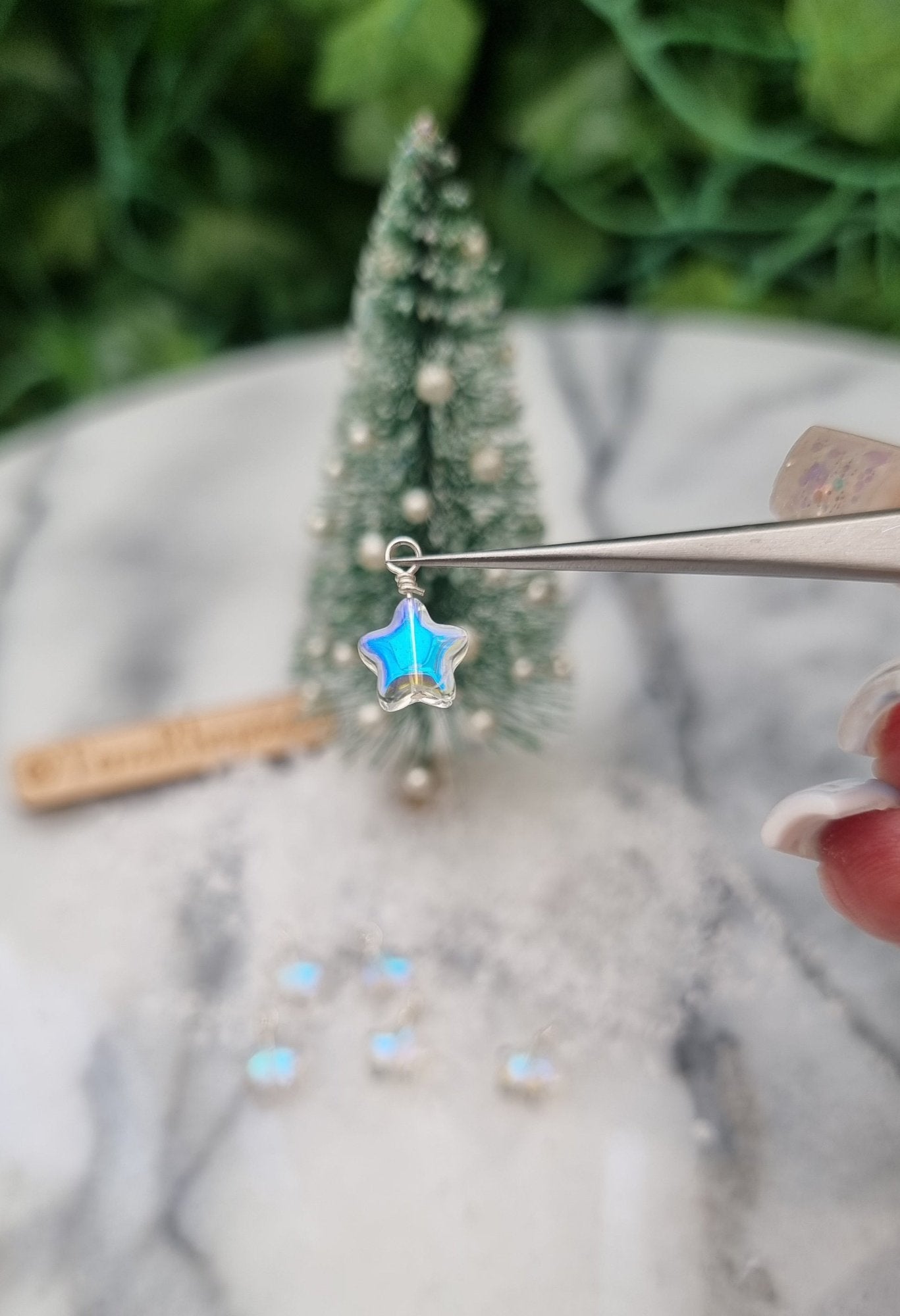 Miniature 1:12 Christmas star baubles is held in tweezers displayed in front of a Christmas tree. The star bauble is a pearlescent blue and clear colour. More star decoration sit at the foot of the tree in the background.@TerraRhapsody is written on wood in the background