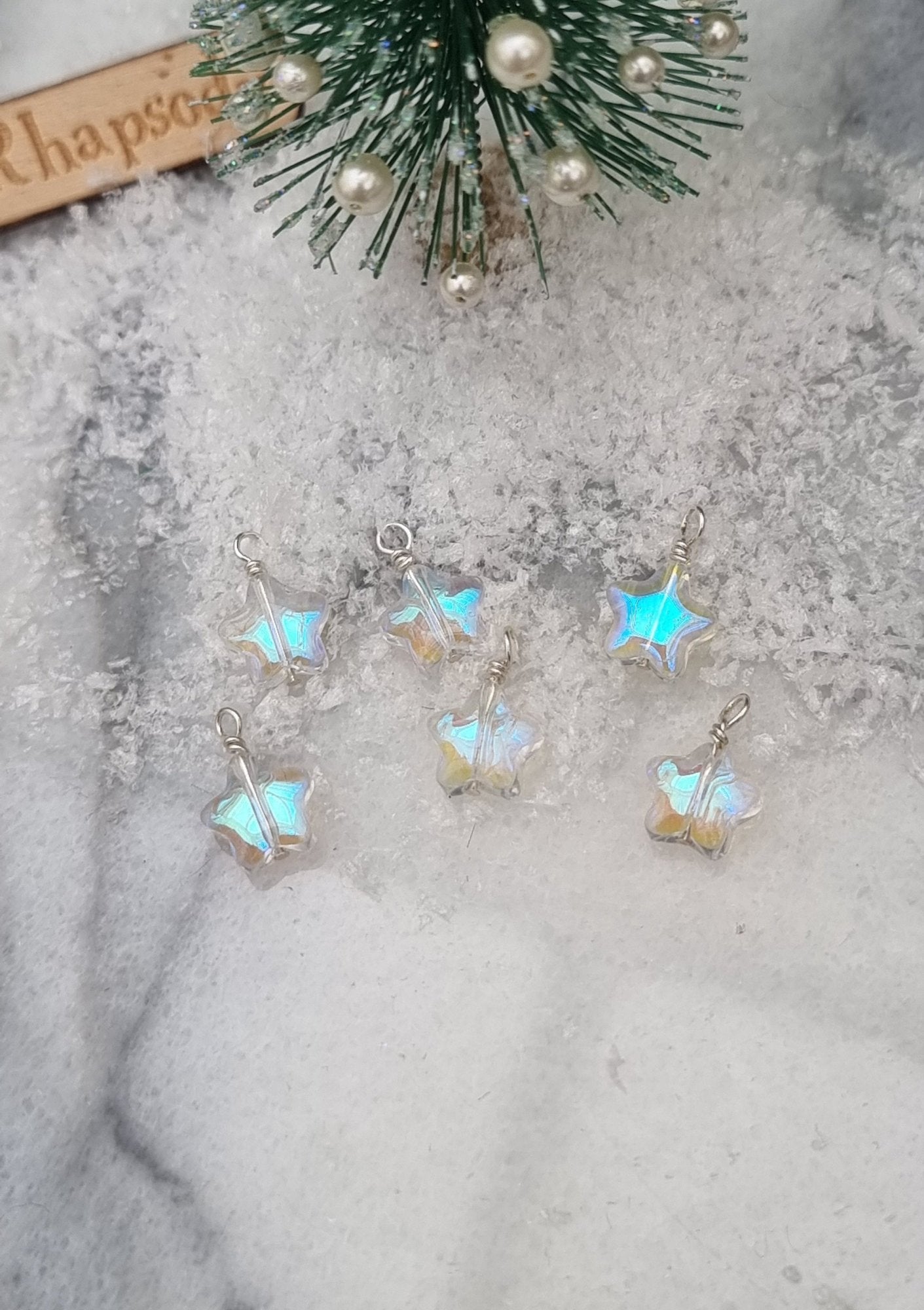 6 Miniature 1:12 Christmas star baubles are displayed on snow at the foot of a miniature Christmas tree. The baubles are star shaped with a pearlescent blue and clear colour. @TerraRhapsody is written on wood in the background