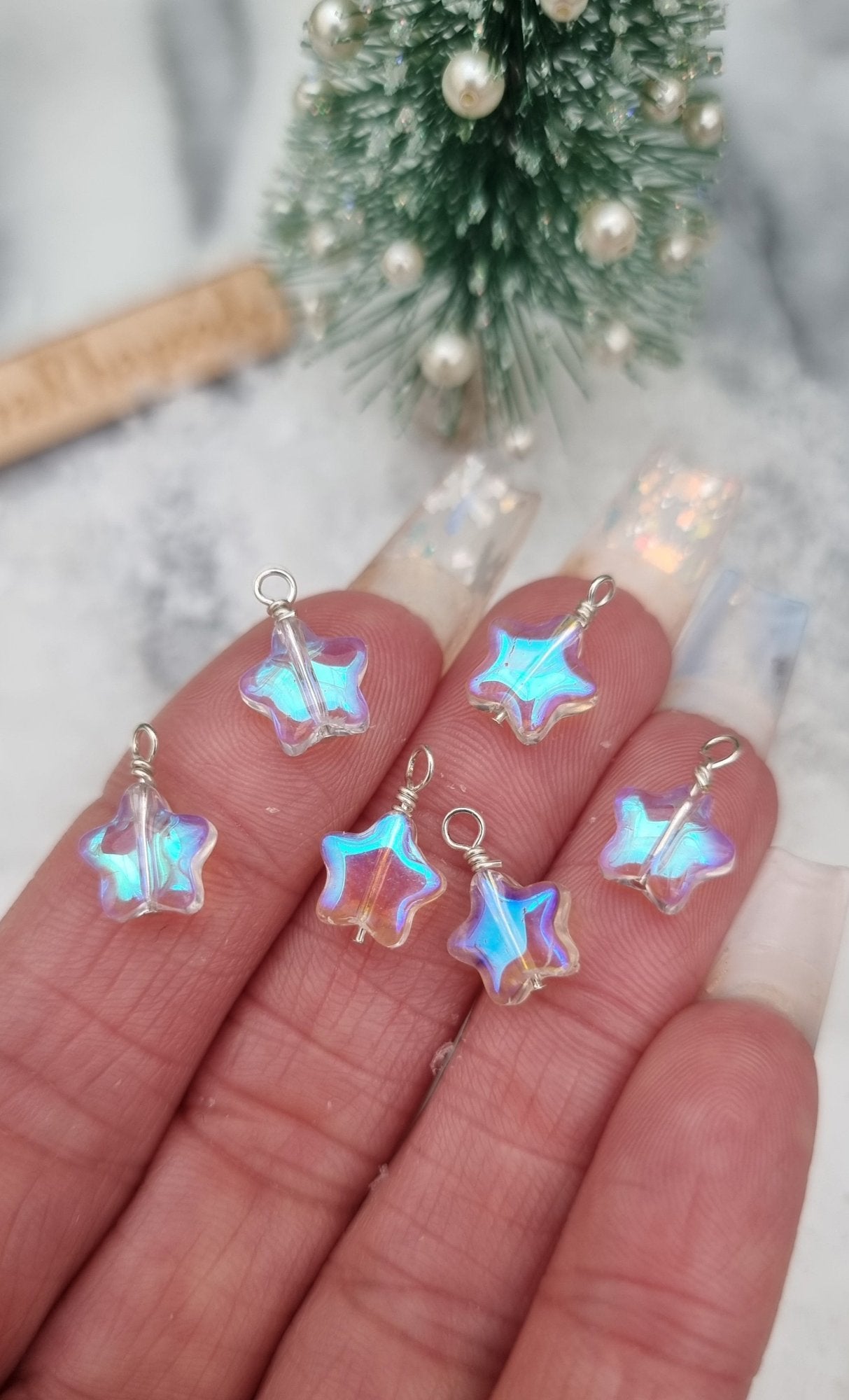 6 Miniature 1:12 Christmas star baubles are displayed in an open hand in front of a miniature Christmas tree. The baubles are star shaped with a pearlescent blue and clear colour. @TerraRhapsody is written on wood in the background