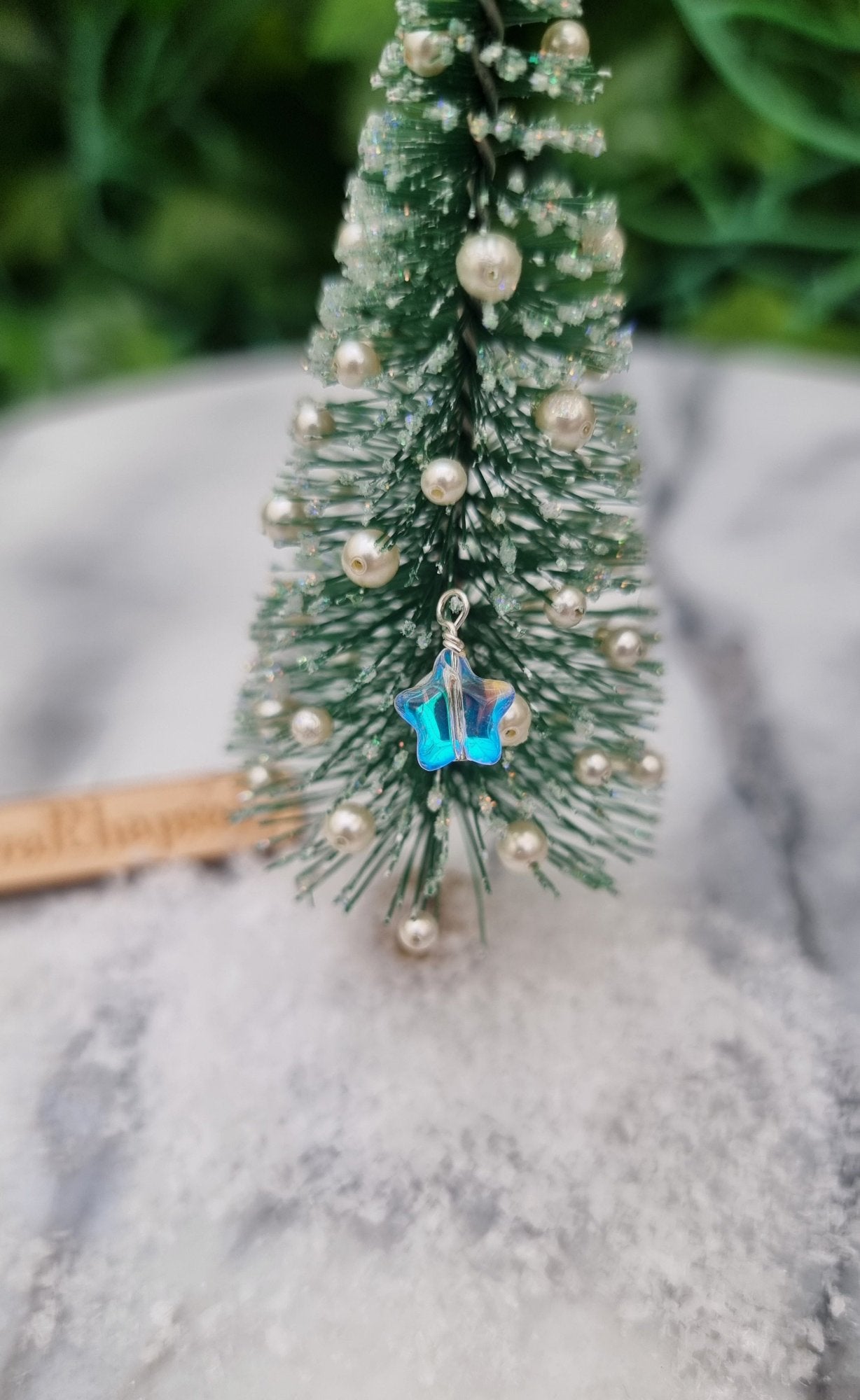 Miniature 1:12 Christmas star bauble is displayed hanging on a miniature Christmas tree. The star bauble is a pearlescent blue and clear colour. @TerraRhapsody is written on wood in the background