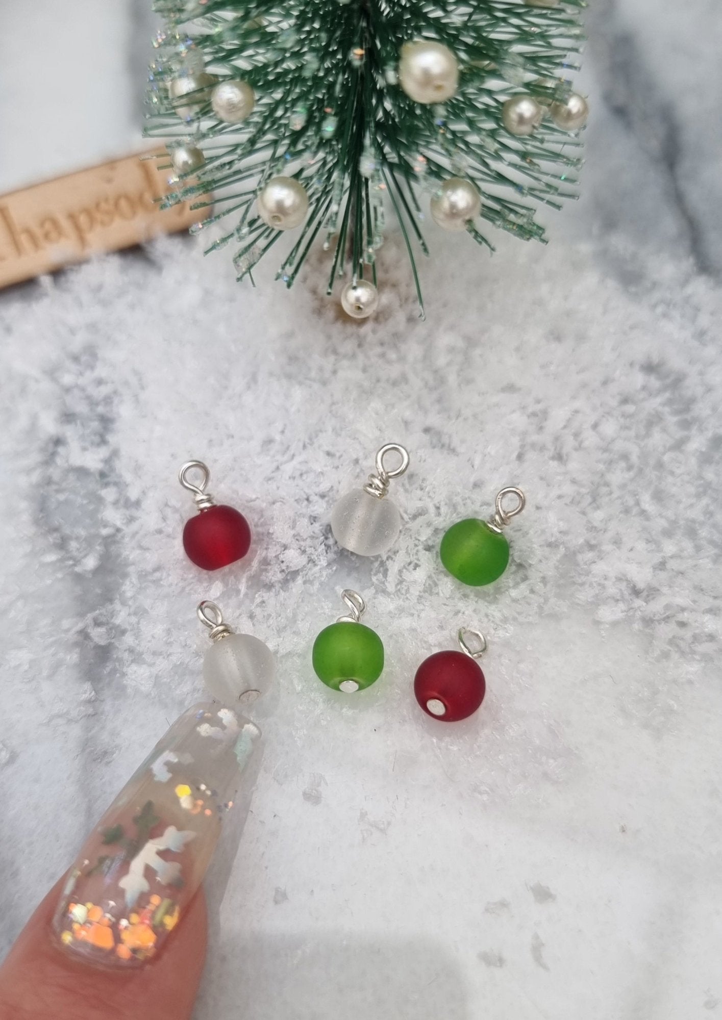 6 Miniature 1:12 Christmas baubles are displayed in front of a Christmas tree. The baubles are round frosted glass in red, green and clear colours. A finger points for scale. @TerraRhapsody is written on wood in the background