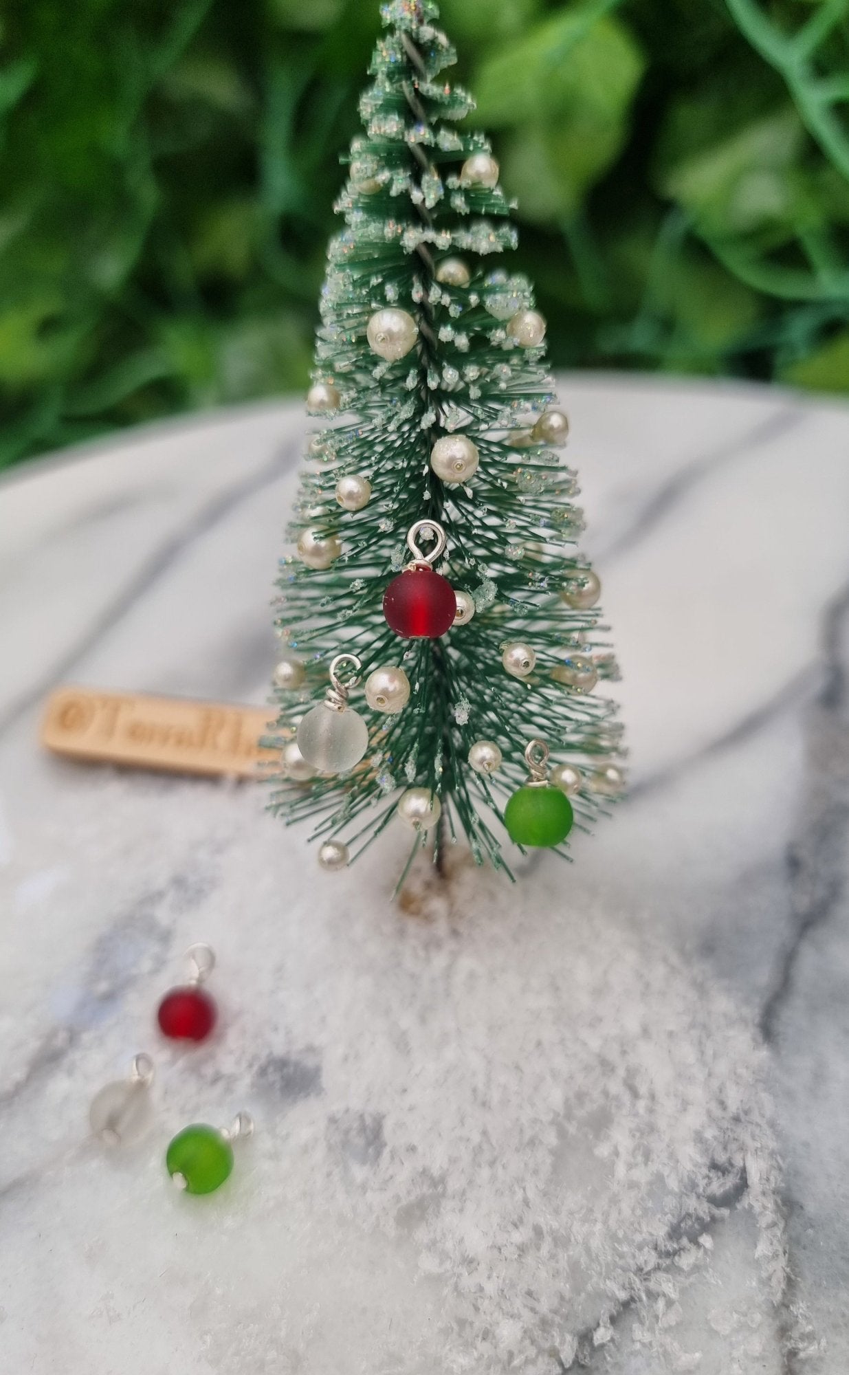 Miniature 1:12 Christmas baubles are displayed on a tiny Christmas tree. The baubles are round frosted glass in red, green and clear colours. More baubles sit at the foot of the tree. @TerraRhapsody is written on wood in the background