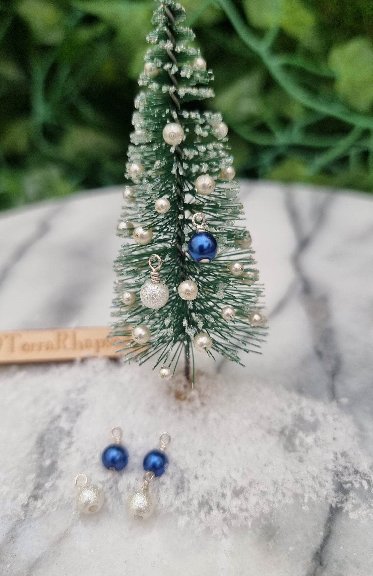 Miniature 1:12 Christmas baubles are displayed on a tiny Christmas tree. The baubles are round in blue and textured white colours. More baubles sit on snow at the foot of the tree. @TerraRhapsody is written on wood in the background