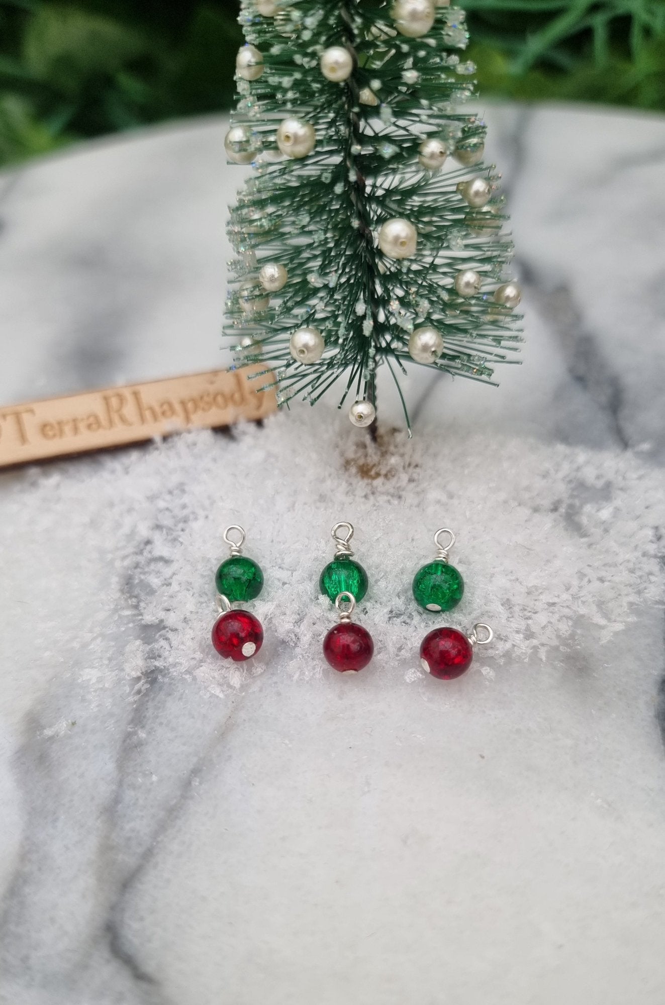 6 Miniature 1:12 Christmas baubles are displayed on snow in front of a Christmas tree. The baubles are crackled glass in bright green and red. @TerraRhapsody is written on wood in the background