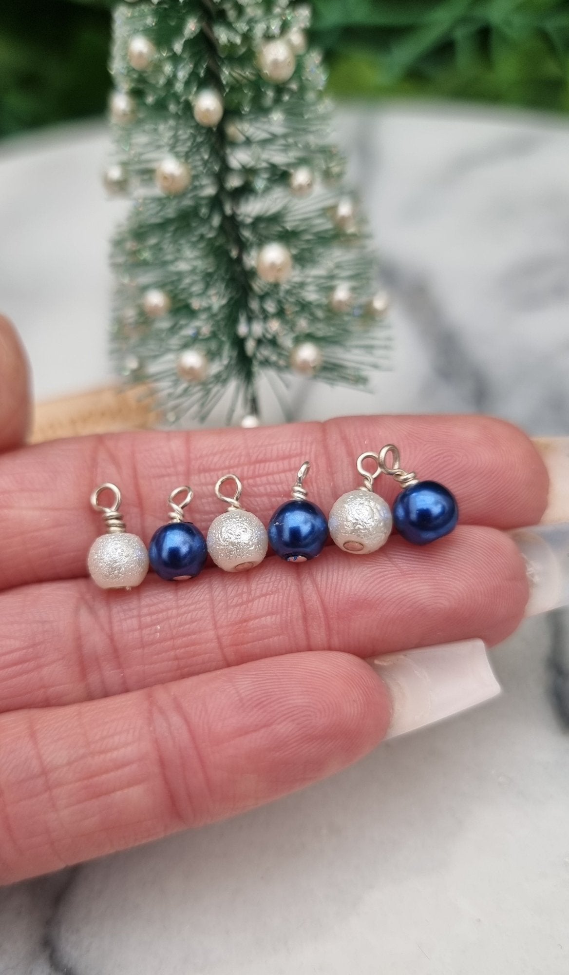 6 Miniature 1:12 Christmas baubles are displayed on a hand front of a Christmas tree. Three baubles are round in pearly blue and three are textured and white in colour. @TerraRhapsody is written on wood in the background.