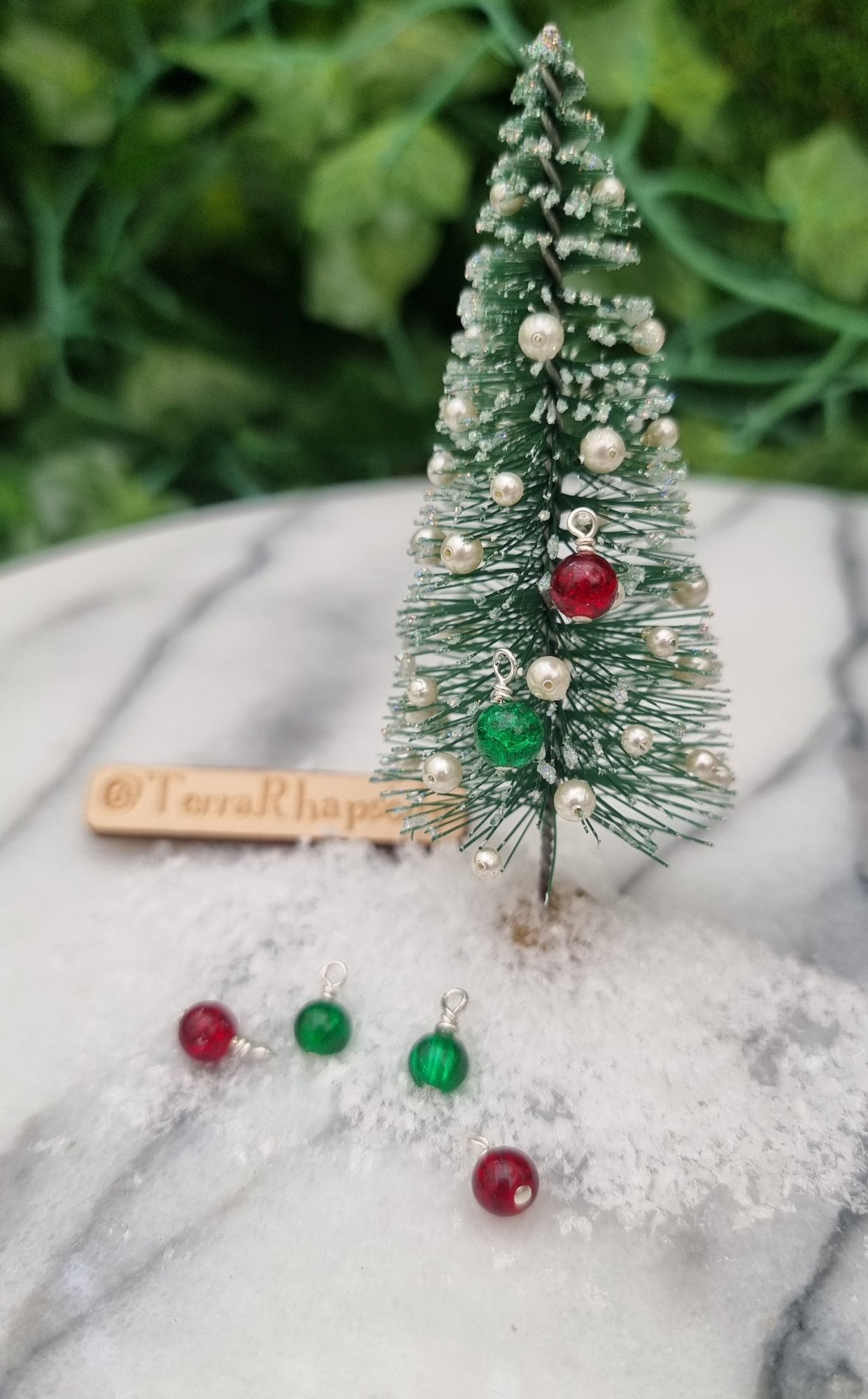 Miniature 1:12 Christmas baubles are displayed on a tiny Christmas tree. The baubles are round crackle glass in bright red and green festive colours.  More baubles sit in snow at the foot of the tree. @TerraRhapsody is written on wood in the background