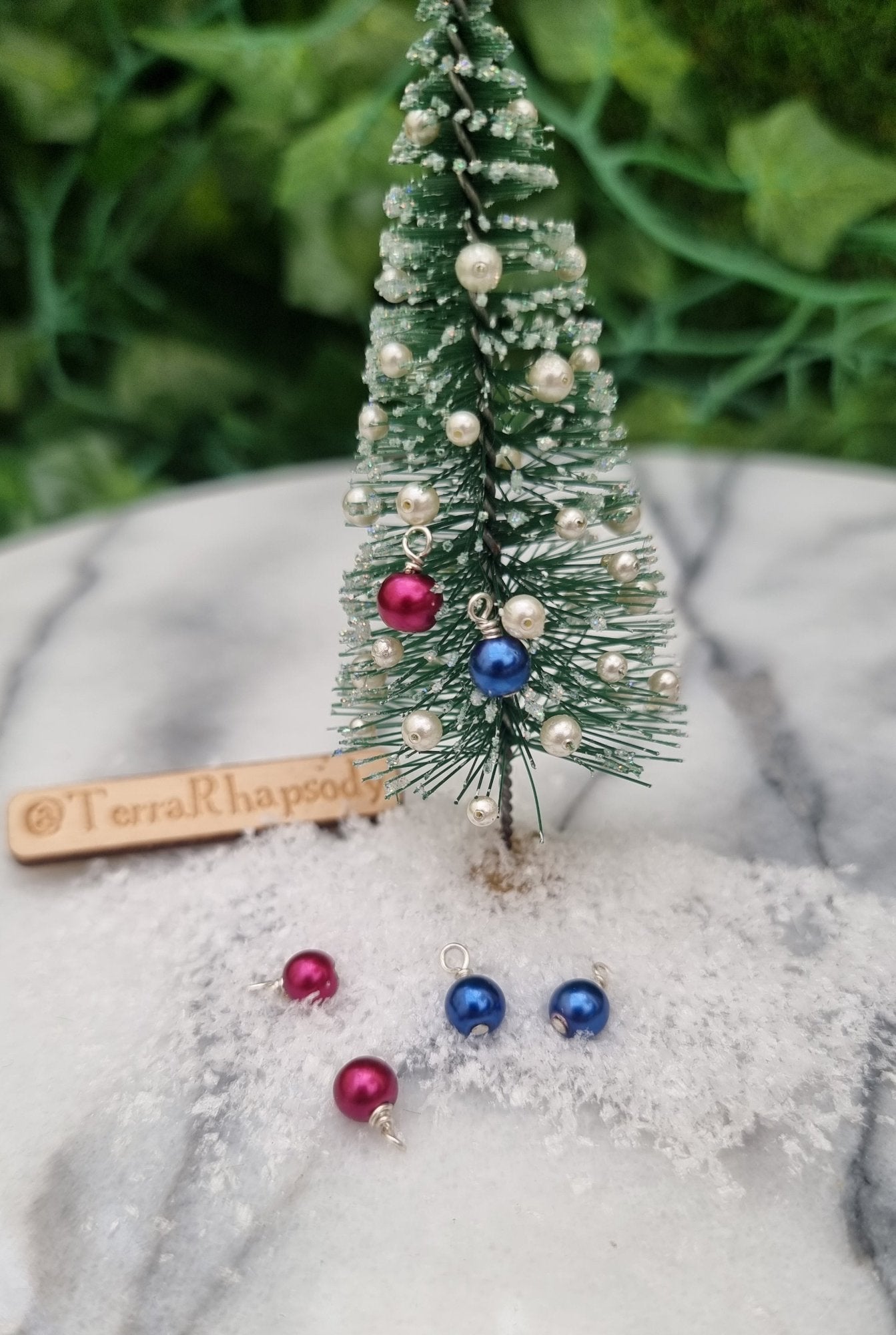 Miniature 1:12 Christmas baubles are displayed on a tiny Christmas tree. The baubles are round in blue and burgundy pearly colours. More baubles sit 9n snow at the foot of the tree. @TerraRhapsody is written on wood in the background