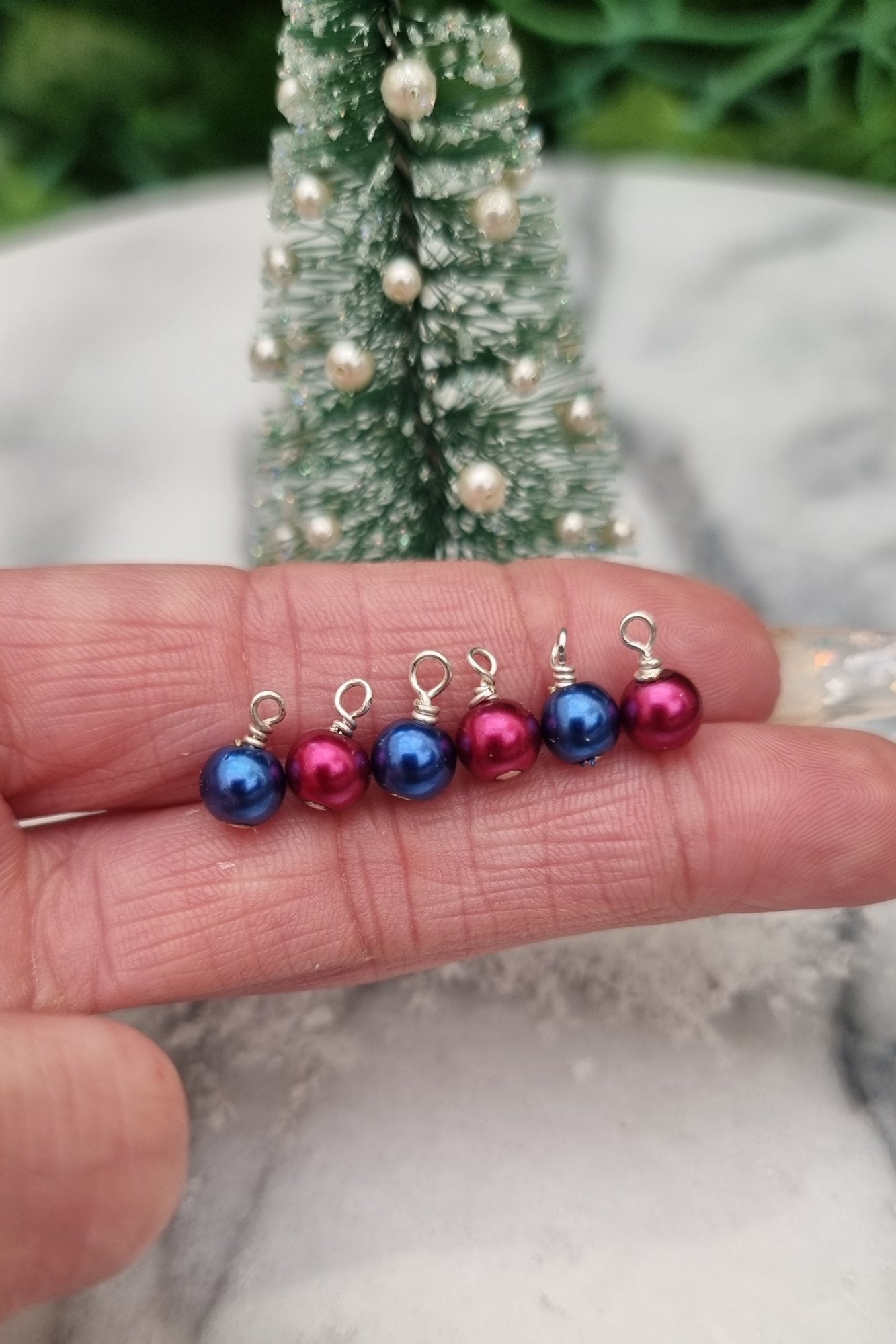 6 Miniature 1:12 Christmas baubles are displayed on a hand front of a miniature Christmas tree. The baubles are round in blue and burgundy colours.