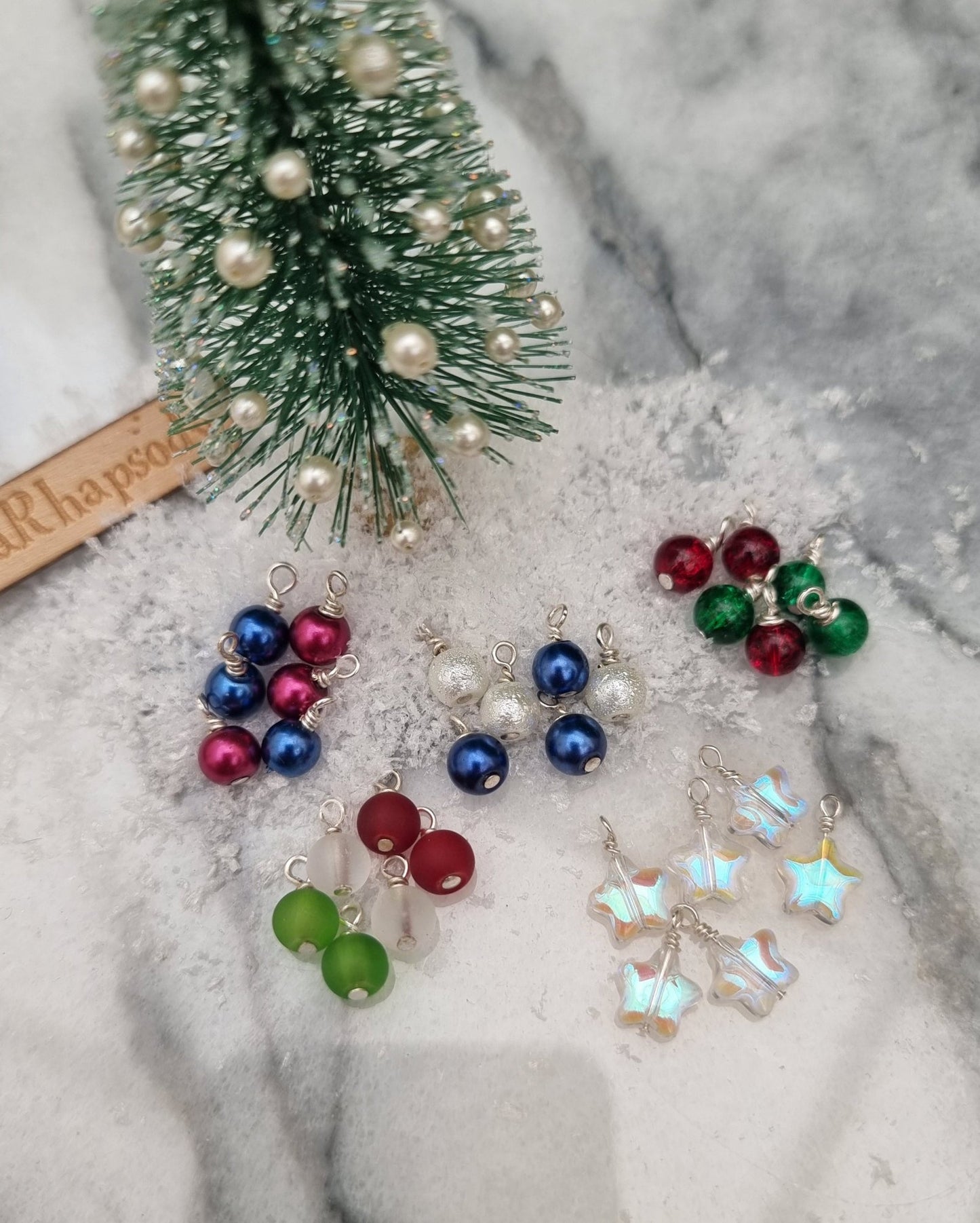 Miniature 1:12 scale Christmas baubles are displayed in front of a tiny tree. The baubles are star shaped and round in different festive colours. @TerraRhapsody is written on wood in the background