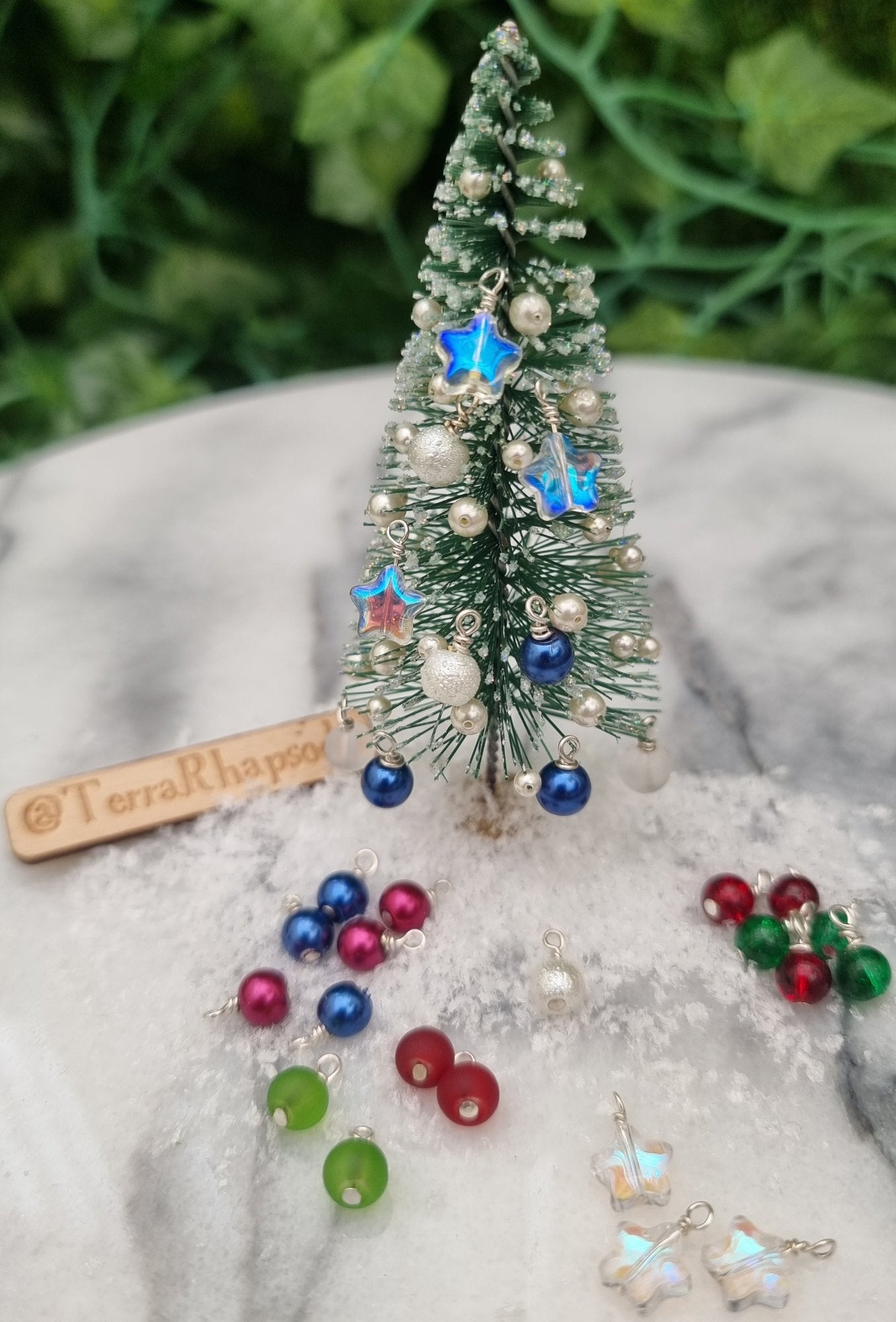 Miniature 1:12 scale Christmas baubles are displayed on a tiny tree. The baubles are star shaped and round in different festive colours with a blue pearlescent coloured decorations hung up. @TerraRhapsody is written on wood in the background