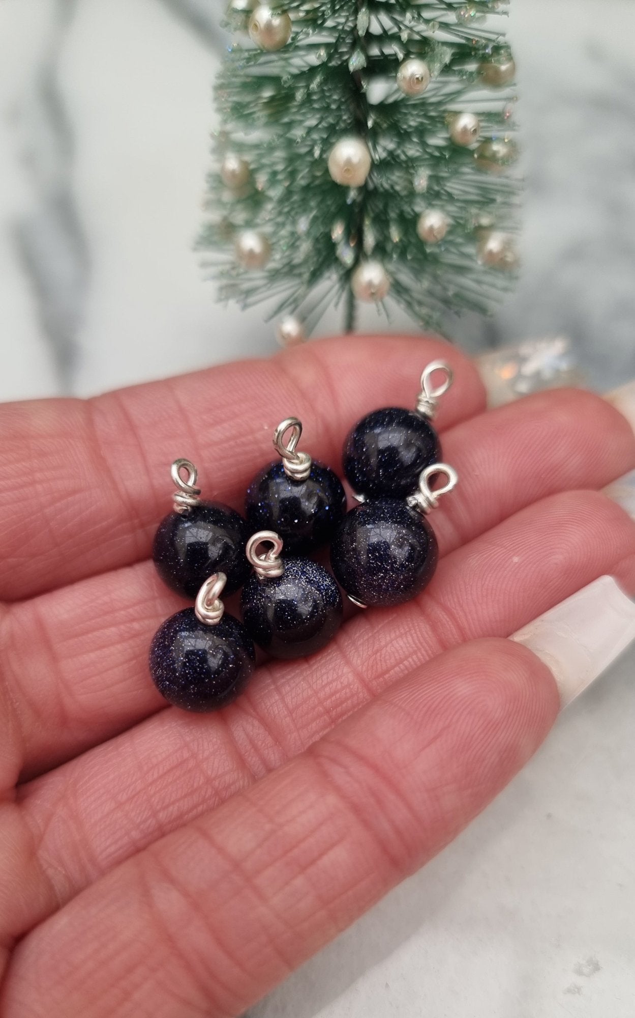 6 Miniature 1:12 Christmas baubles are displayed a hand in front of a Christmas tree. The baubles are glittery blue goldstone. @TerraRhapsody is written on wood in the background