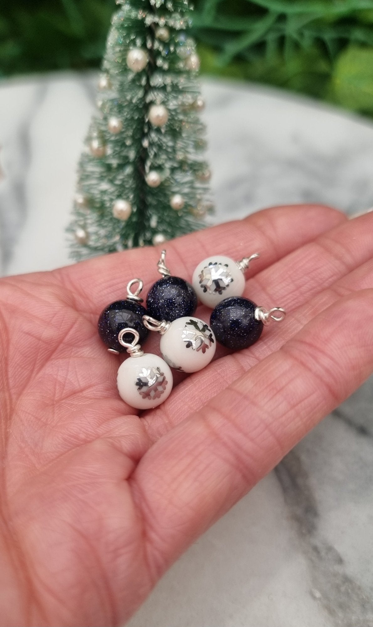 6 Miniature 1:12 Christmas baubles are displayed a hand in front of a Christmas tree. The baubles are white with silver snowflake motifs and glittery blue goldstone. @TerraRhapsody is written on wood in the background