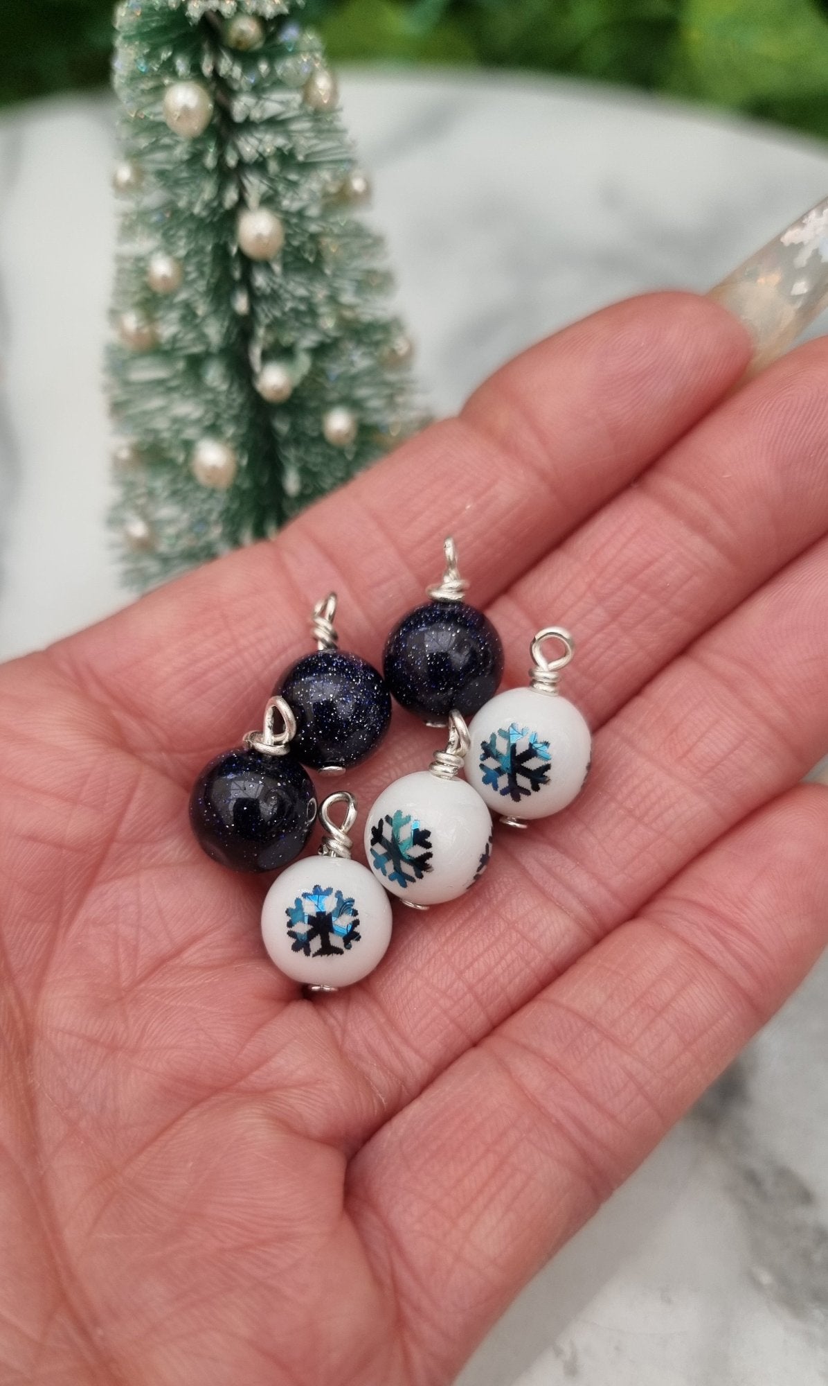 6 Miniature 1:12 Christmas baubles are displayed a hand in front of a Christmas tree. The baubles are white with blue snowflake motifs and glittery blue goldstone. @TerraRhapsody is written on wood in the background