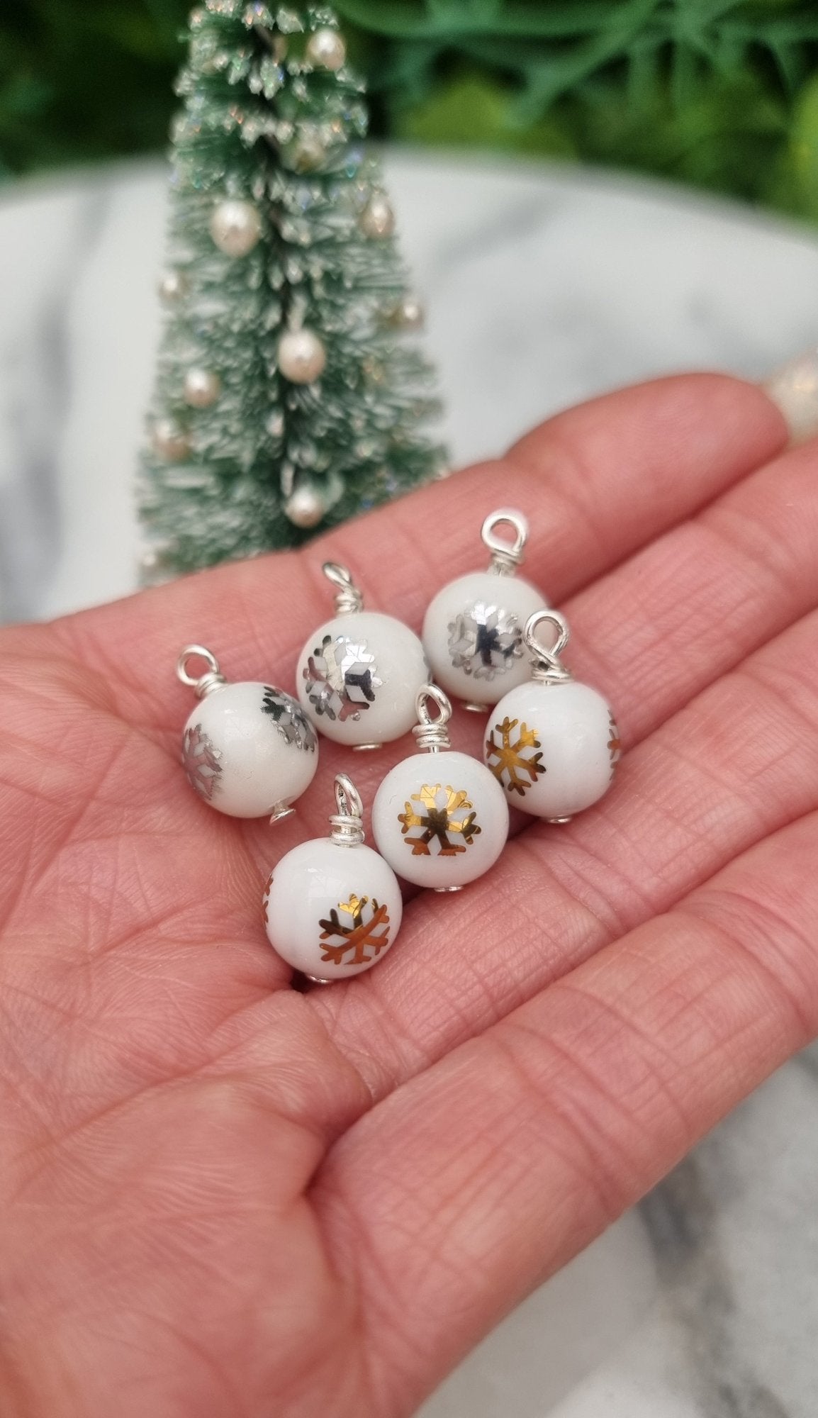 6 Miniature 1:12 Christmas baubles are displayed a hand in front of a Christmas tree. The baubles are white with silver and gold snowflake and Christmas tree motifs. @TerraRhapsody is written on wood in the background