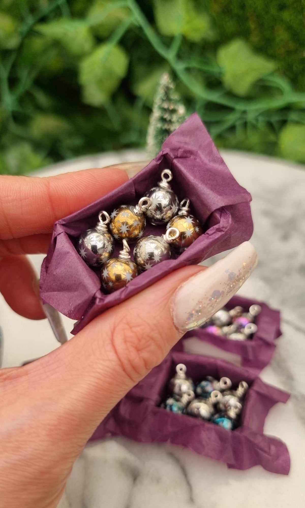 Miniature 1:12 scale open tray box contains 6 Christmas baubles. The baubles are silver and gold coloured with white star motifs. The box is lined with purple paper, displayed between two fingers in front of a Christmas tree