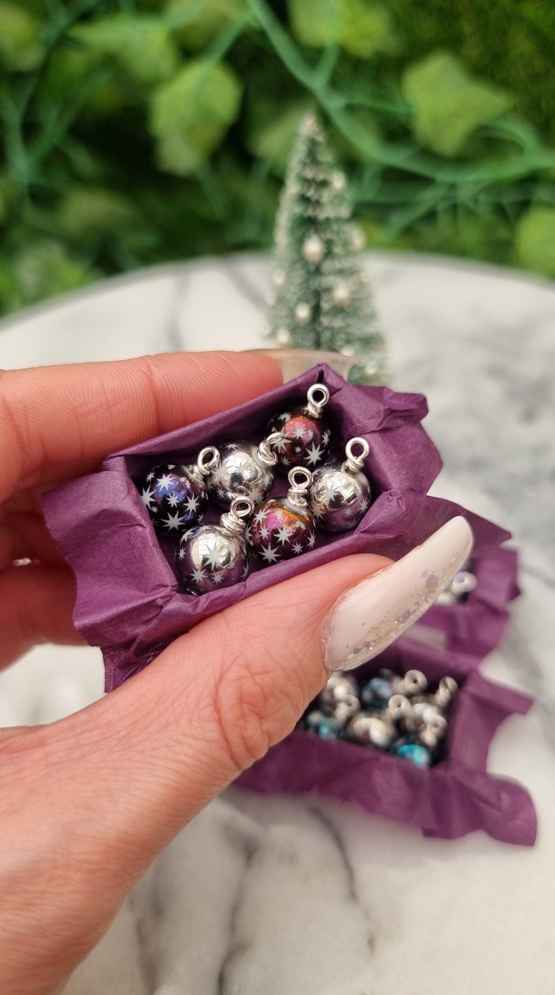 Miniature 1:12 scale open tray box contains 6 Christmas baubles. The baubles are silver and metallic purple coloured with white star motifs. The box is lined with purple paper, displayed between two fingers in front of a Christmas tree