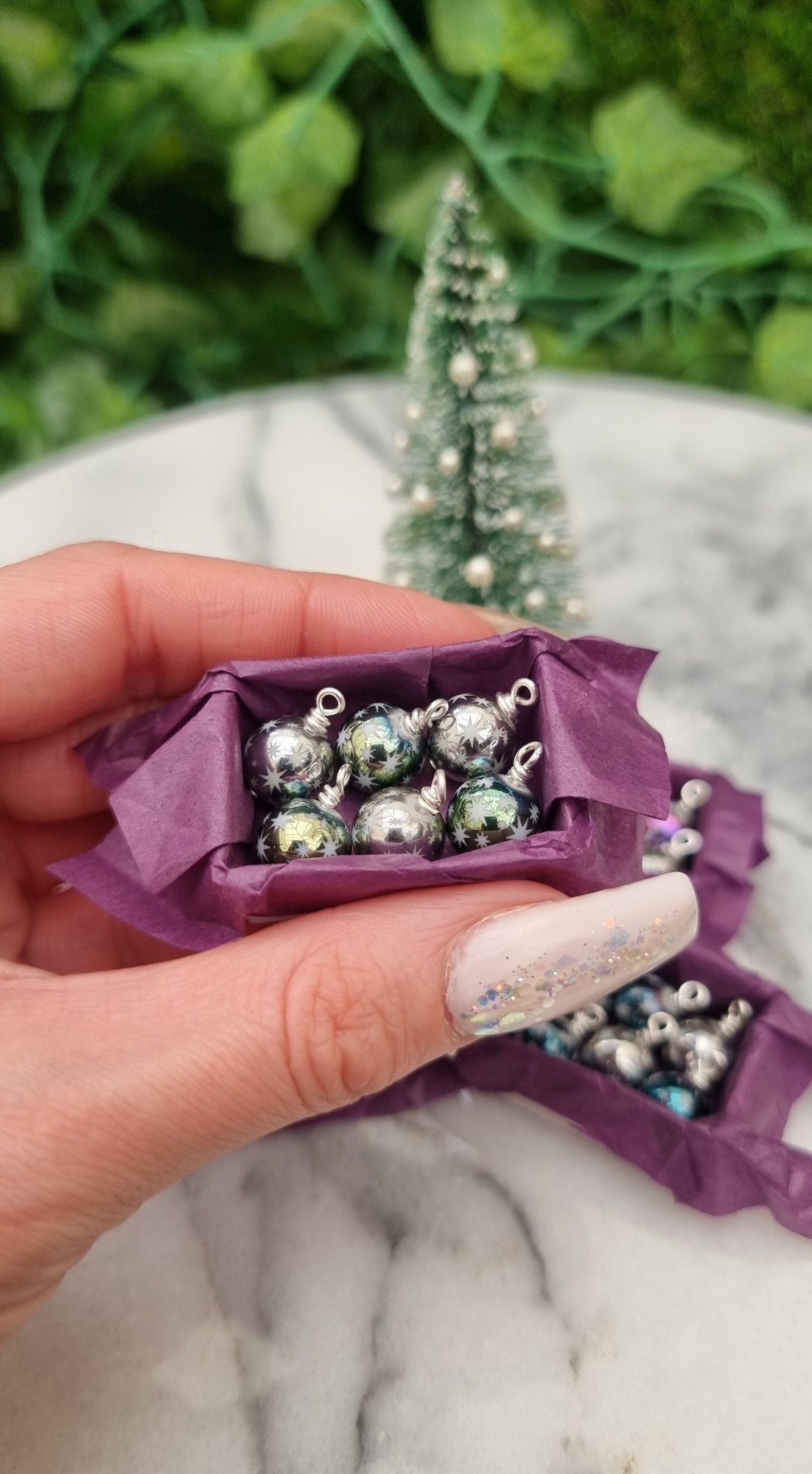 Miniature 1:12 scale open tray box contains 6 Christmas baubles. The baubles are silver and pastel green coloured with white star motifs. The box is lined with purple paper, displayed between two fingers in front of a Christmas tree